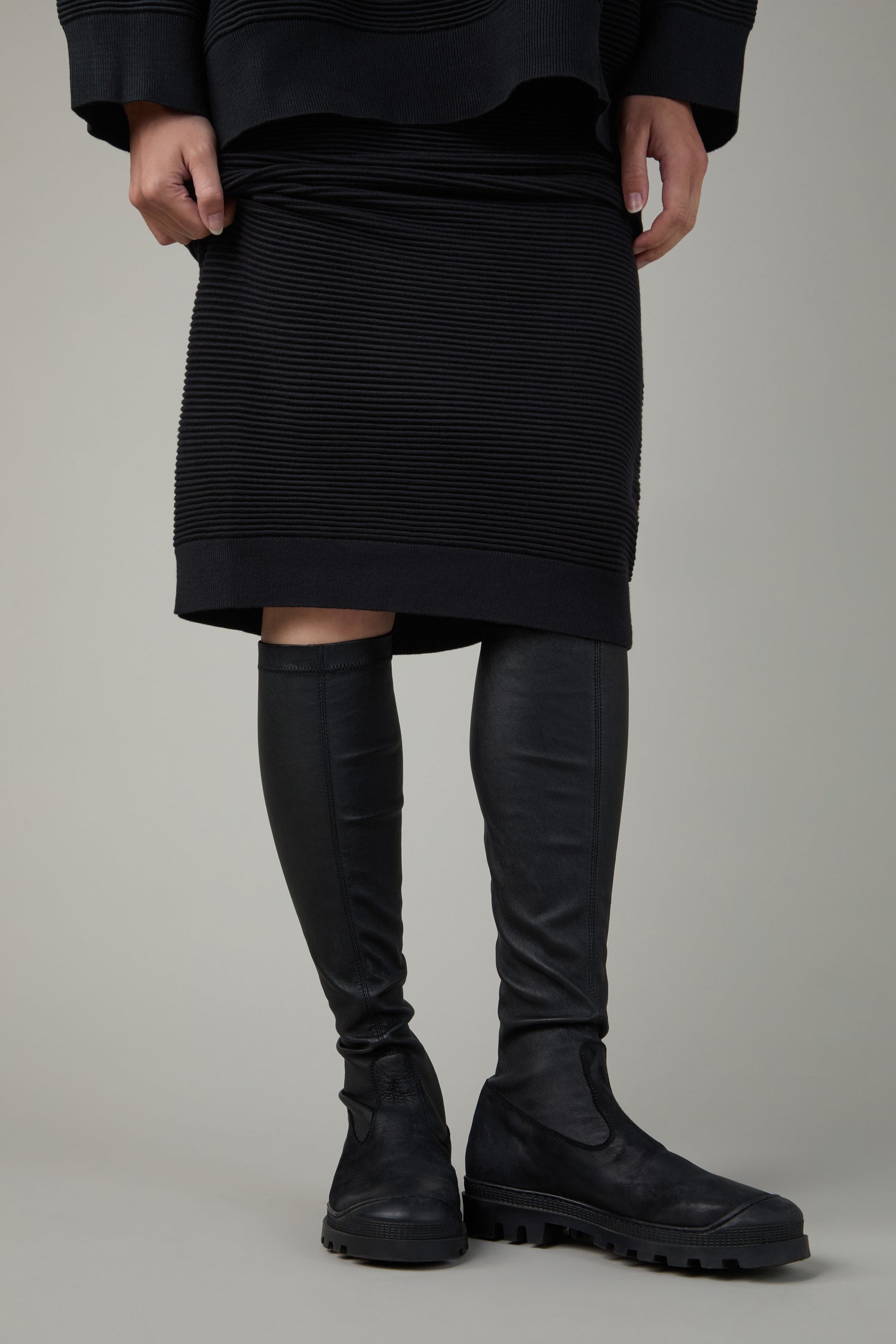 A person wearing a black ribbed skirt, a black top, and Stretch Long Nappa Leather Boots stands against a plain background.