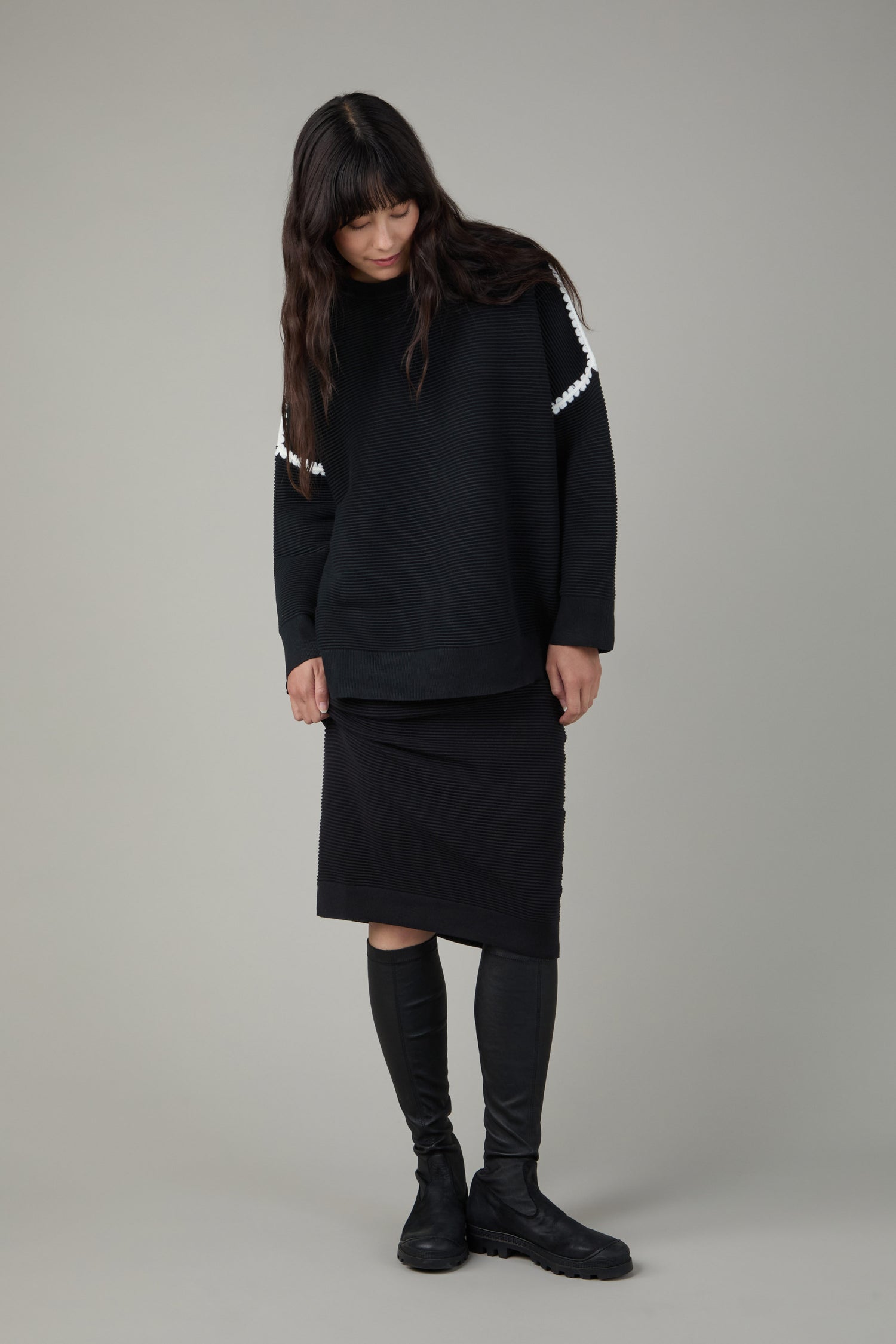 A person with long dark hair wearing a black sweater with white trim, black skirt, and Stretch Long Nappa Leather Boots stands against a plain gray background.