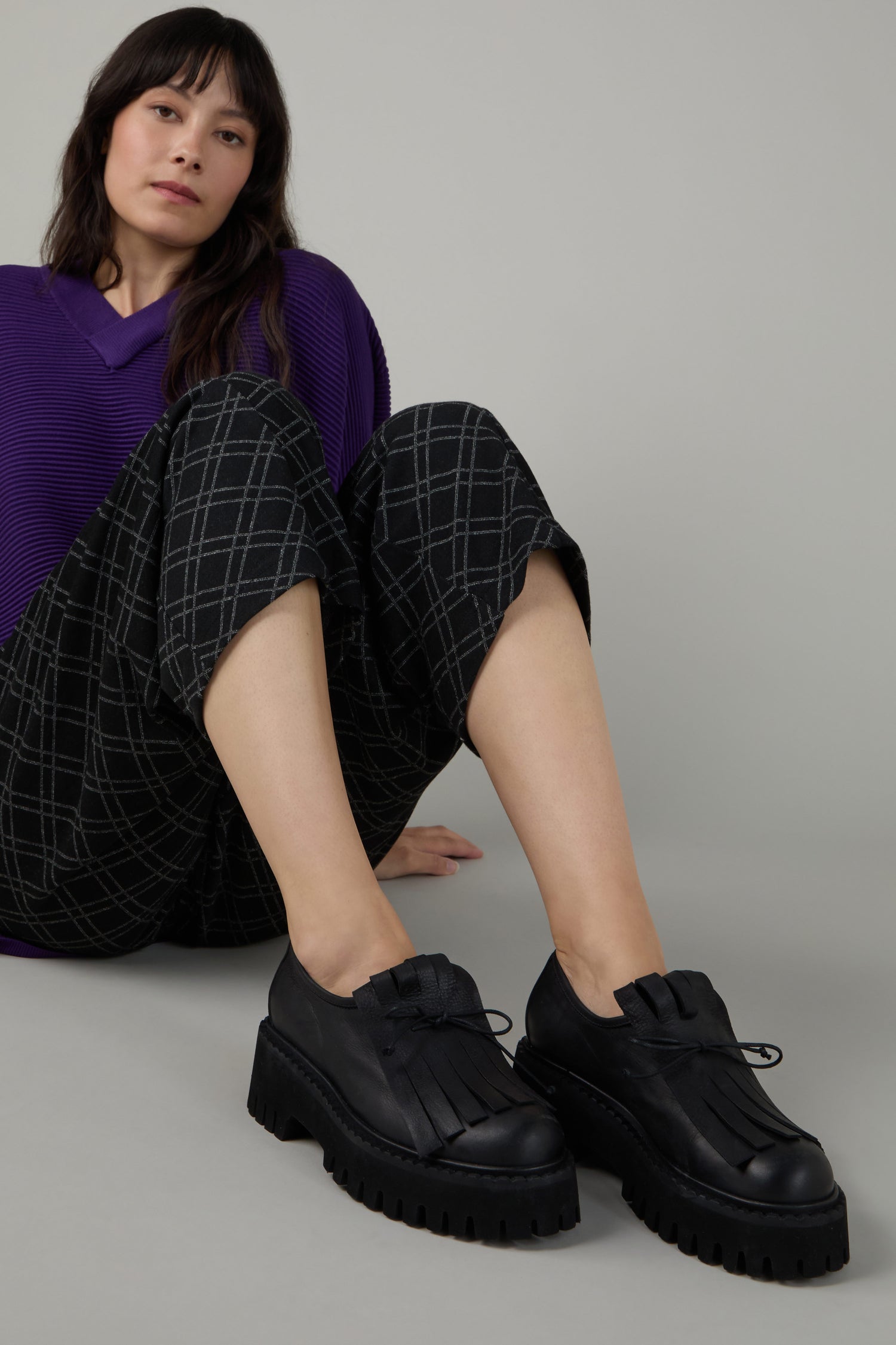 Person sitting on the floor wearing a purple top, black plaid pants, and Fringe Top Leather Brogues—black handcrafted chunky lace-up shoes with stylish fringe detail.