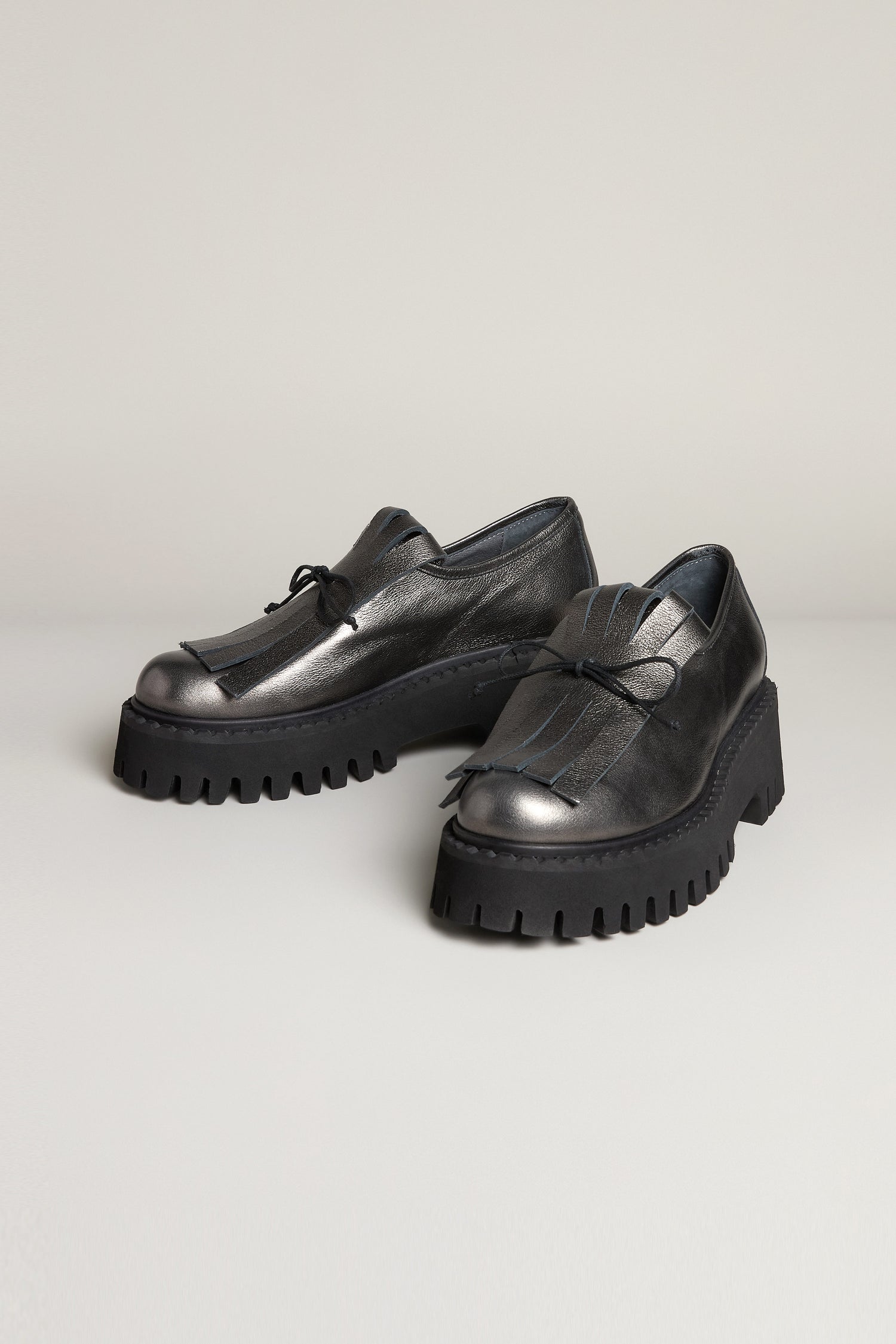 Fringe Top Leather Brogues—black chunky-soled handcrafted leather shoes with a metallic finish and decorative laces—displayed on a light-colored surface.