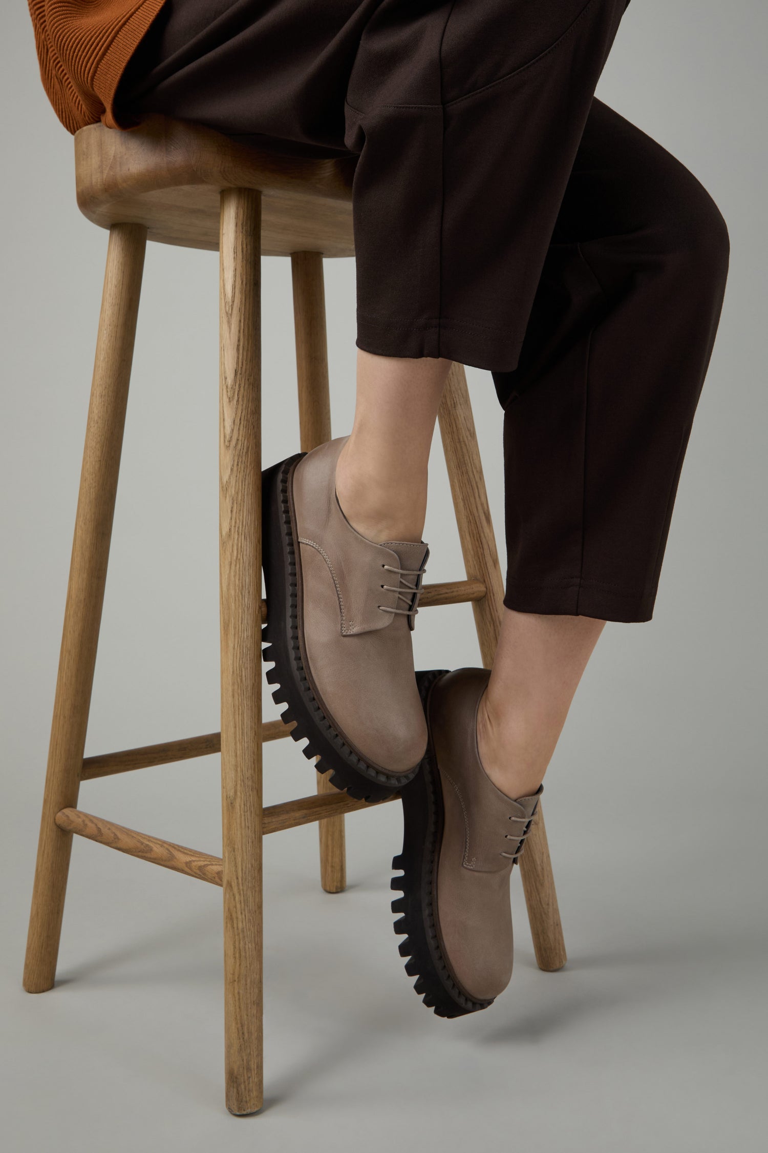A person sits on a wooden stool, wearing brown pants and Two Tone Lace Up Leather Brogues with contemporary design and thick black soles.