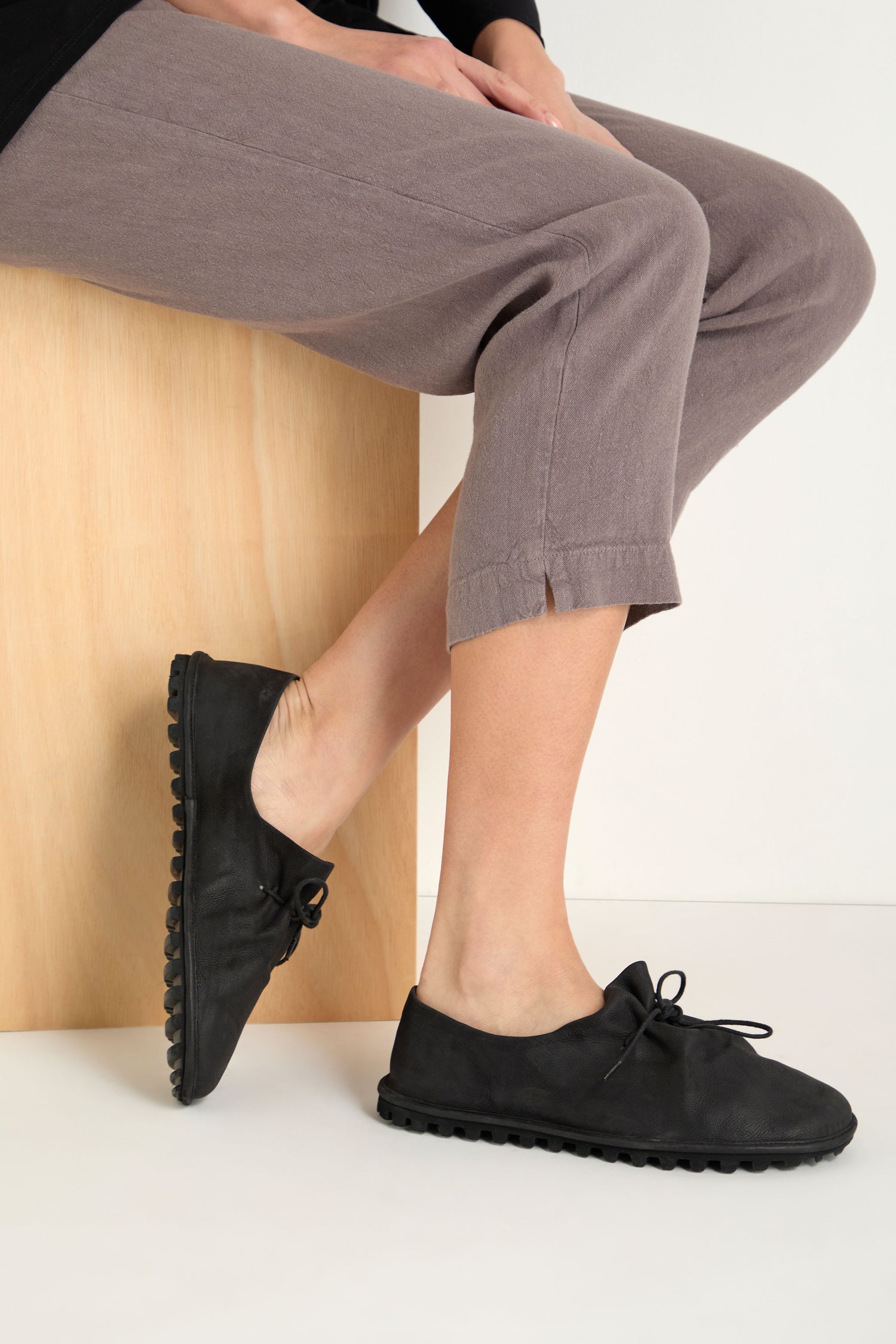 A person in cropped mauve pants and Crushed Leather Lace Up Shoes, handmade in Italy, sits on a light wooden bench against a plain background.