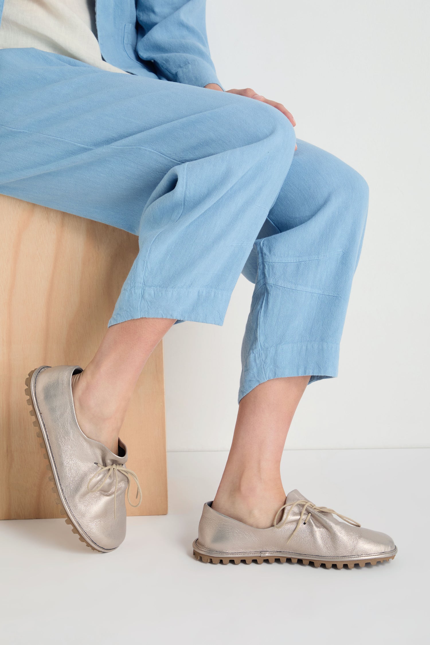 A person sits on a light wooden bench wearing light blue pants and Crushed Leather Lace Up Shoes, handmade in Italy, featuring stylish brown soles.