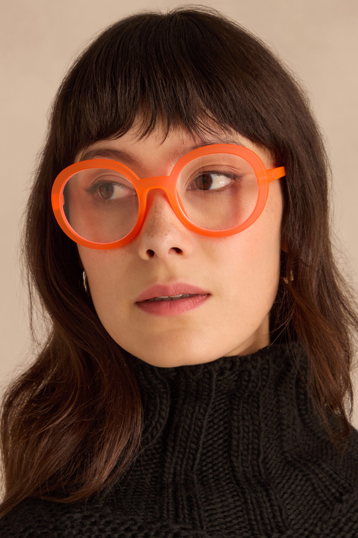 Woman with medium-length brown hair wears Neon Peach Round Reading Glasses and a black turtleneck, looking slightly to the side.
