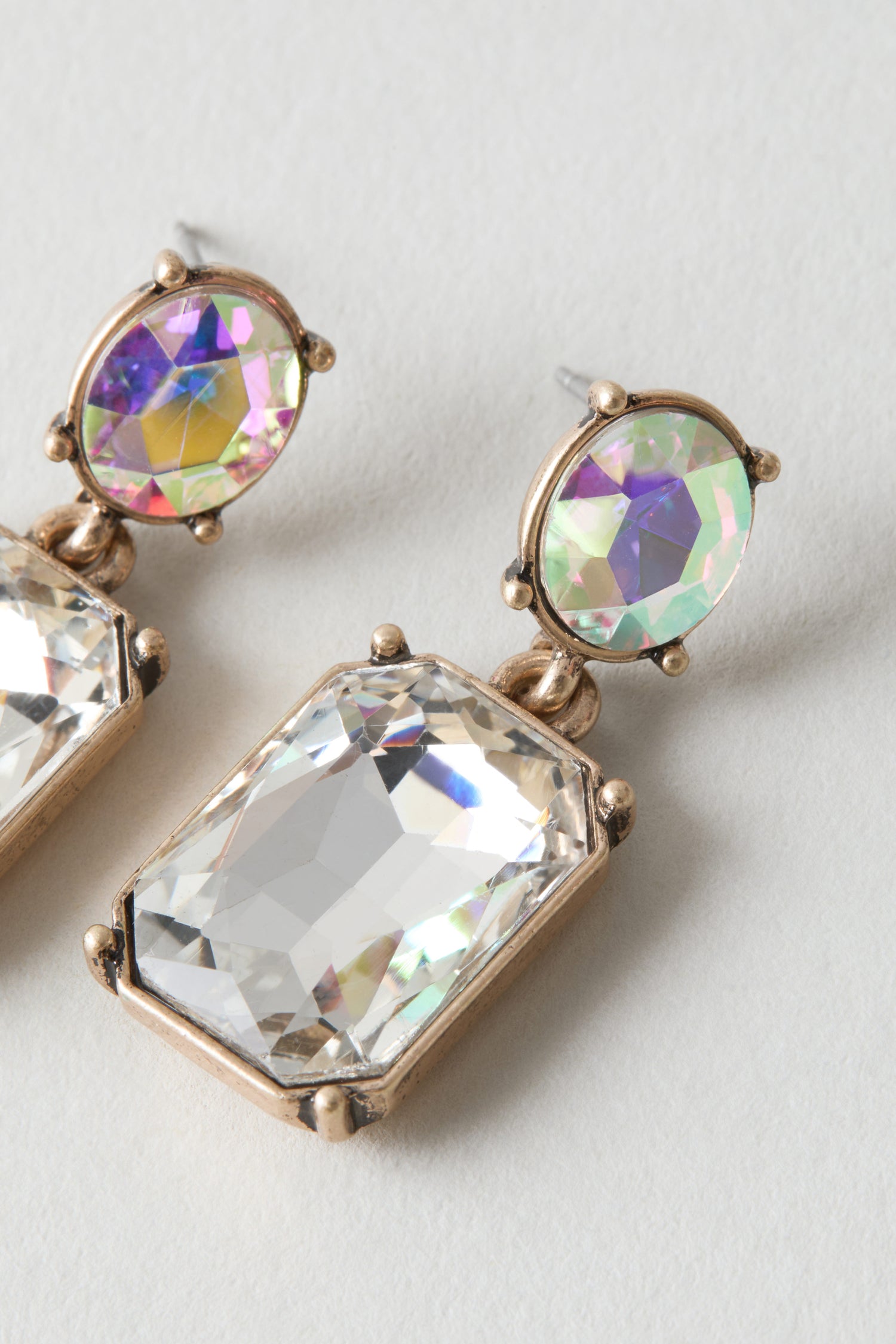 Close-up of Twin Gemstone Earrings: each earring showcases a round iridescent stone atop a large rectangular clear gem, both set in gold-tone metal, displayed on a light background.