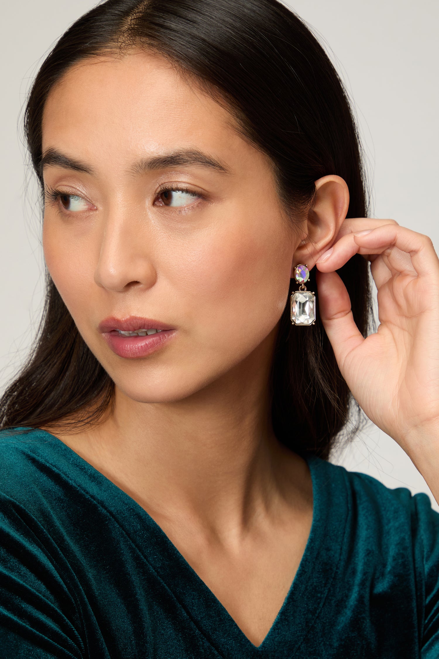 A woman with straight dark hair in a green velvet top shows off Twin Gemstone Earrings—striking statement earrings with a large rectangular clear gemstone and a smaller blue stone above it.