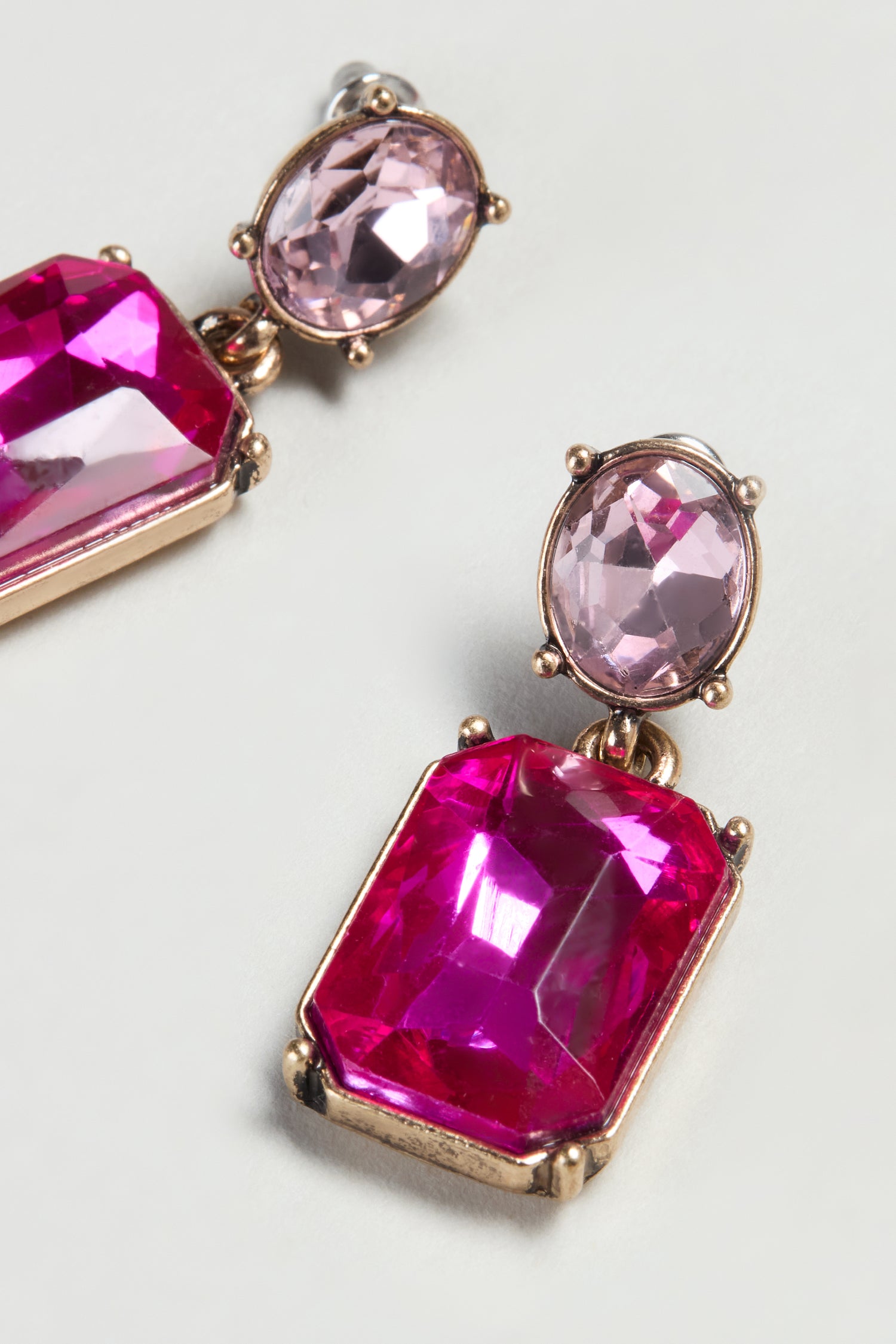 The Twin Gemstone Earrings are gold-tone statement earrings with large rectangular pink gemstones and smaller oval light purple stones, set on a light background in a modern design.