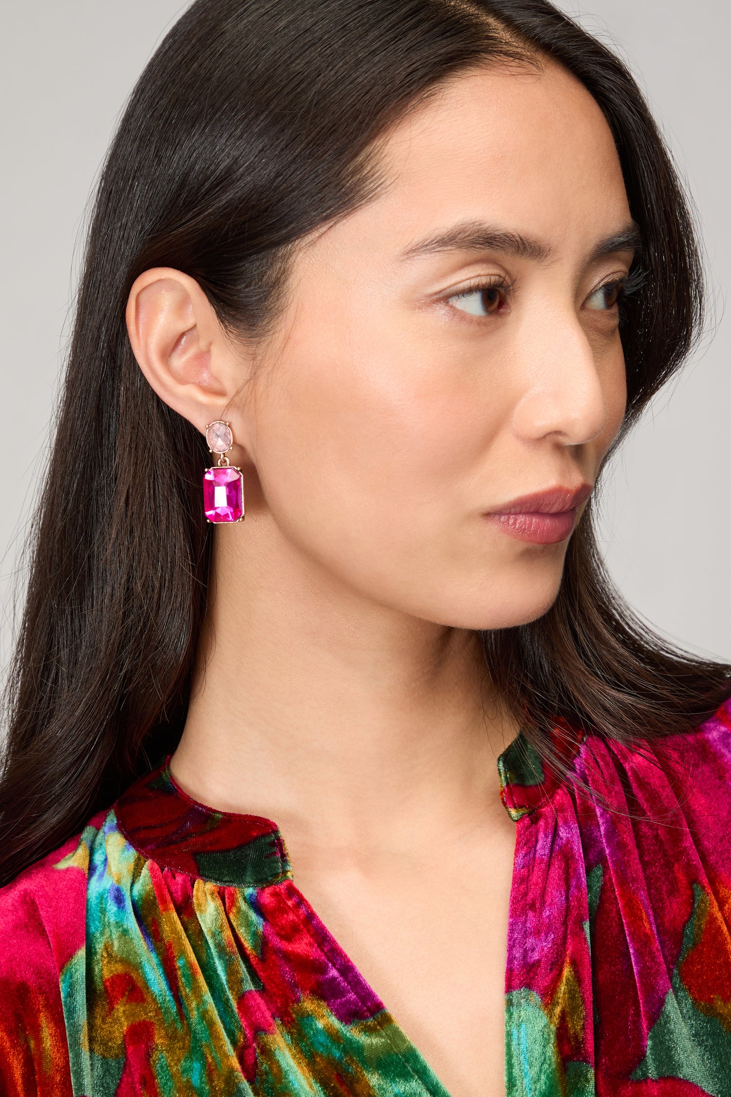 A woman with long dark hair wears a vibrant multicolored velvet top and striking Twin Gemstone Earrings featuring large pink stones, gazing to the side.