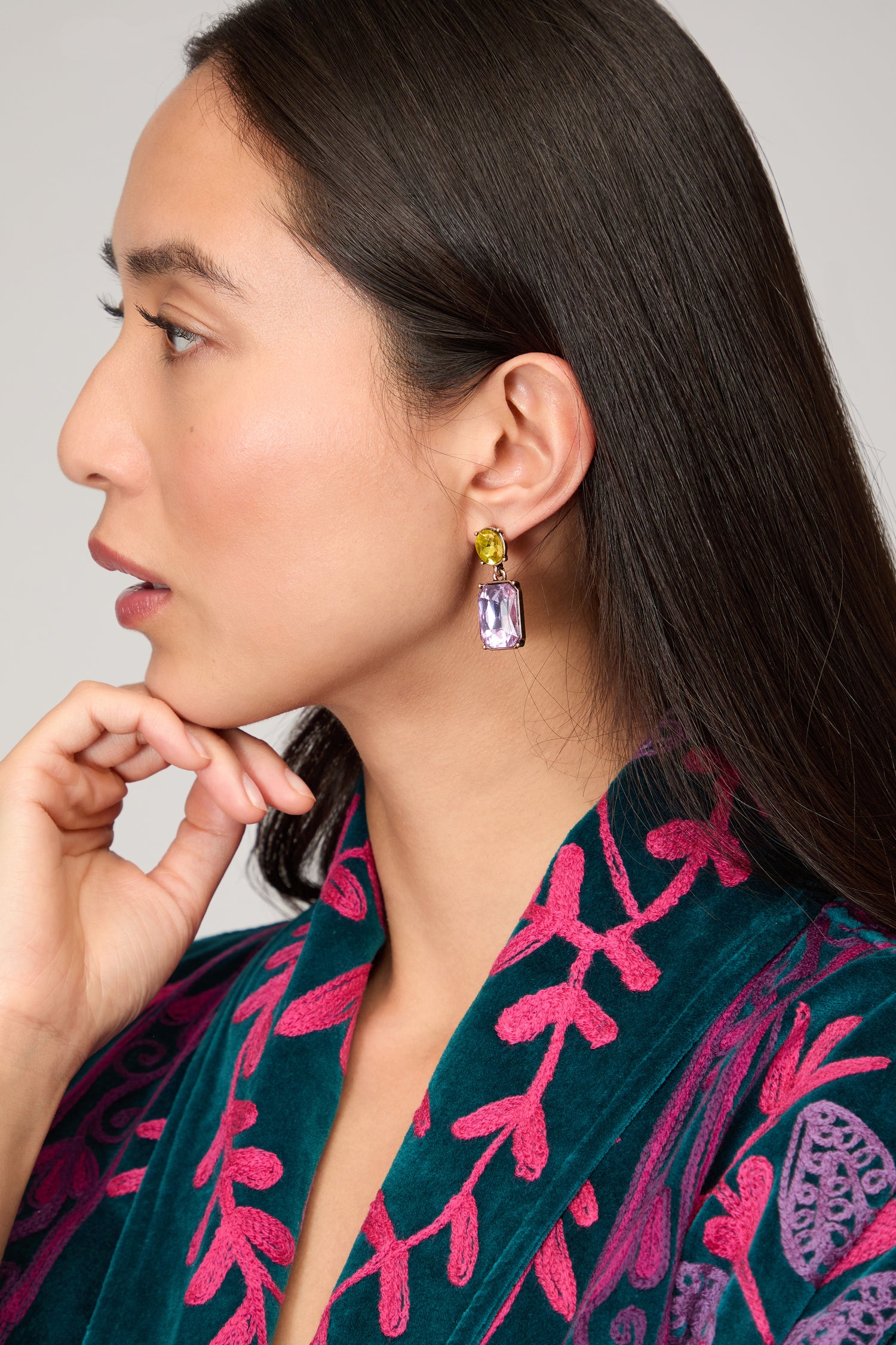 A woman in a blue and pink patterned robe poses in profile, highlighting the Twin Gemstone Earrings with gold and purple accents as her hand gently touches her chin.