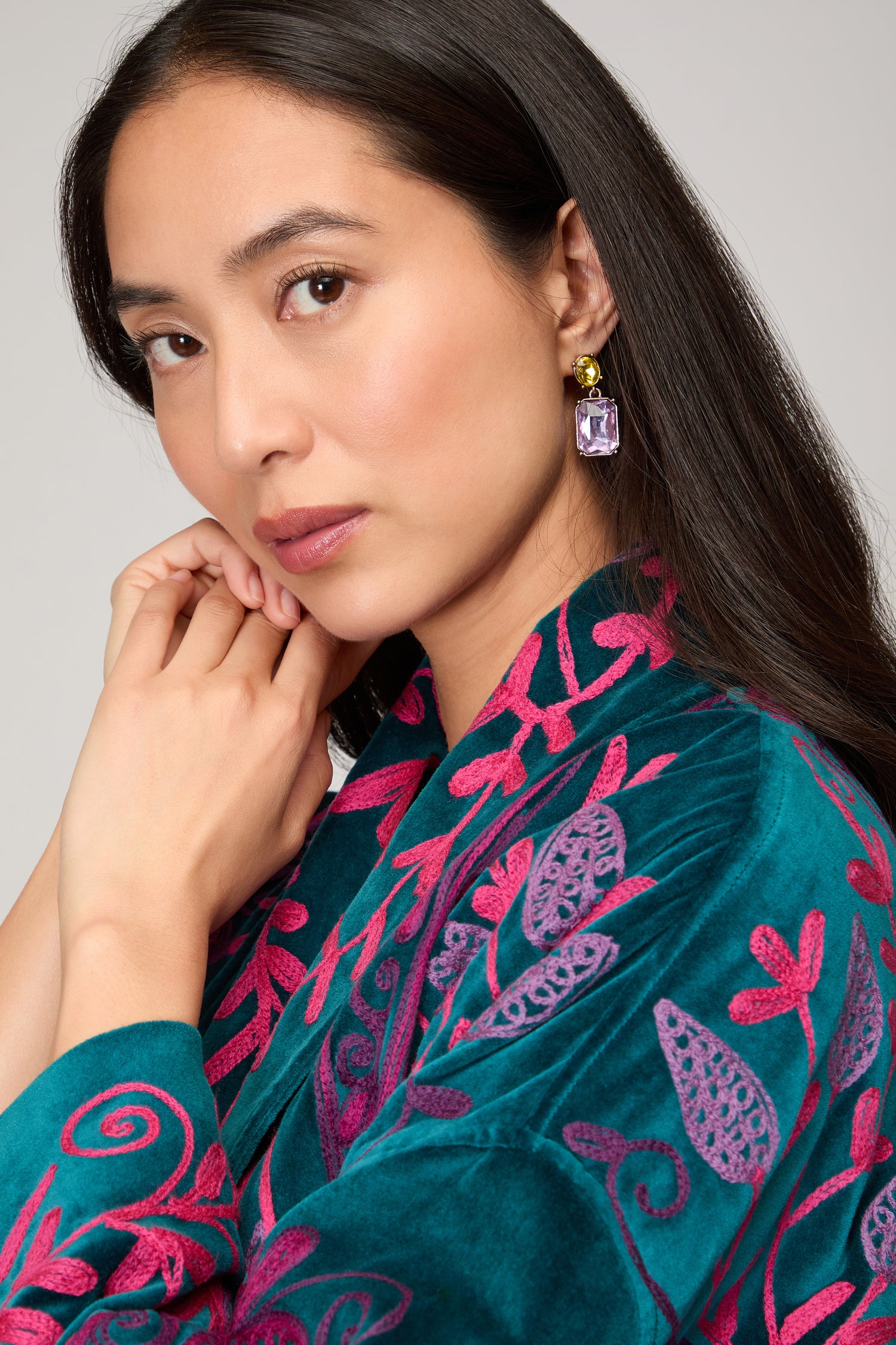 A woman with long dark hair wears Twin Gemstone Earrings and an embroidered teal and magenta garment, gazing calmly at the camera.
