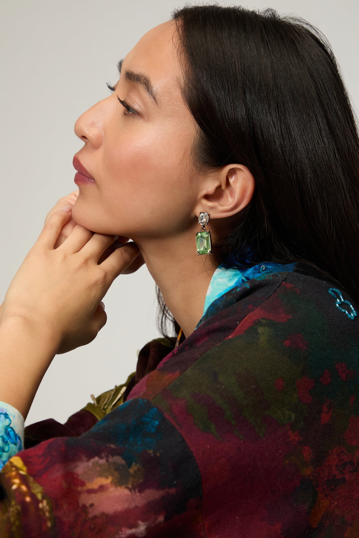 A woman in profile wears Twin Gemstone Earrings, featuring large green stones, paired with a colorful top as she rests her chin on her hand against a neutral background.