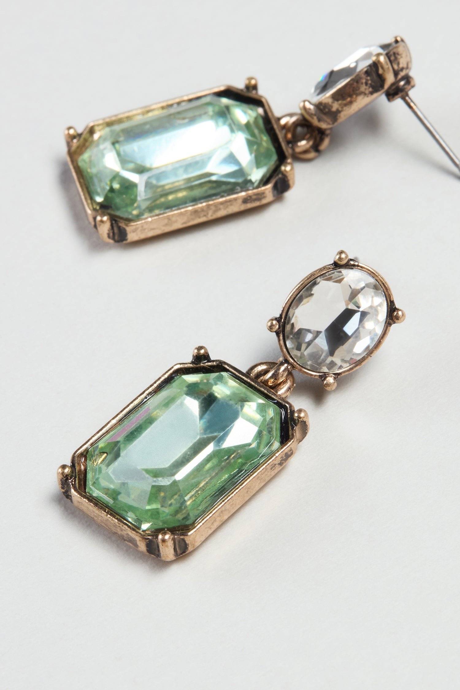 Twin Gemstone Earrings with a modern design feature gold settings, green rectangular and clear oval stones, shown on a light gray background.