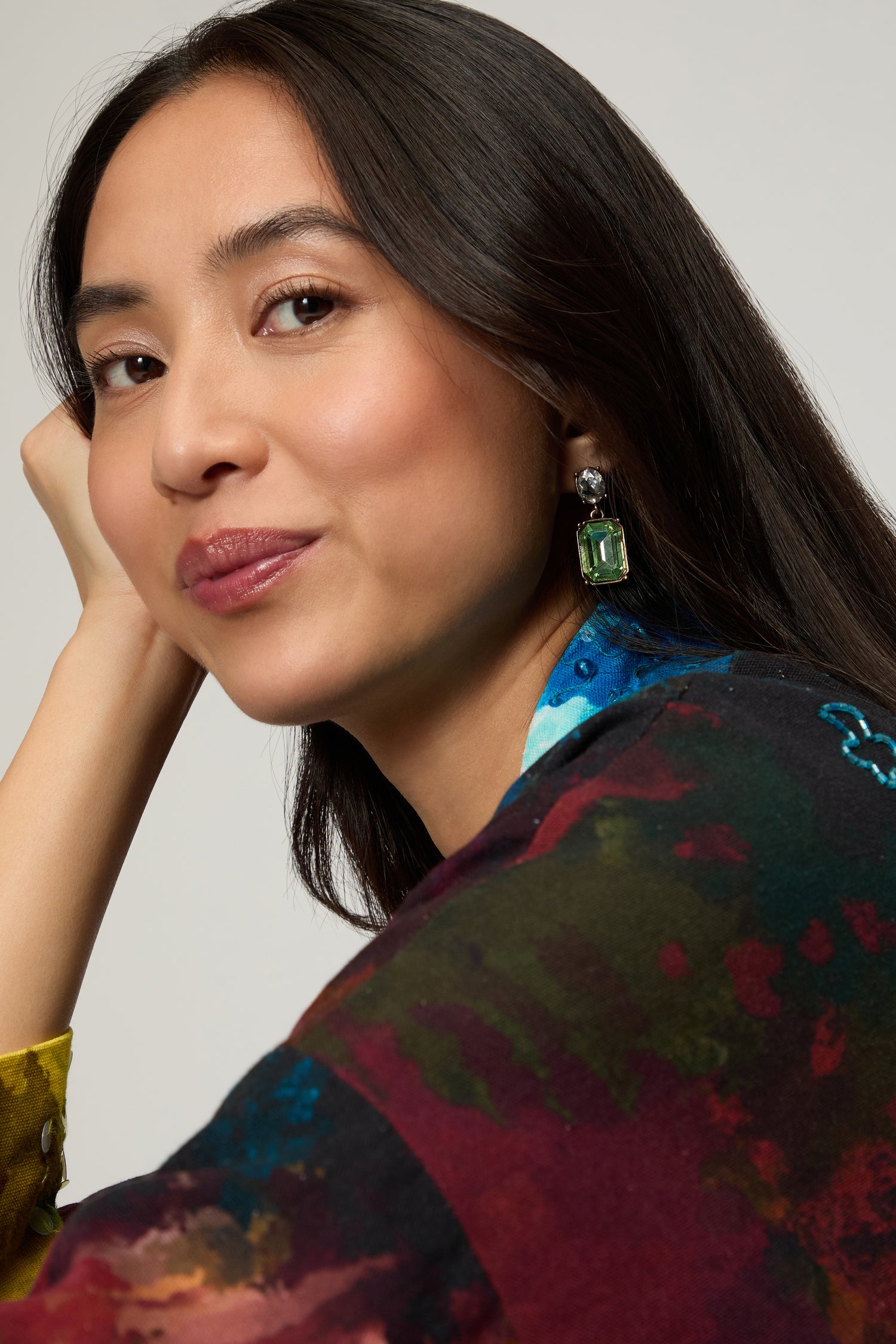A woman with long dark hair, wearing a colorful top and the Twin Gemstone Earrings featuring green stones, poses with her hand on her face while looking at the camera.