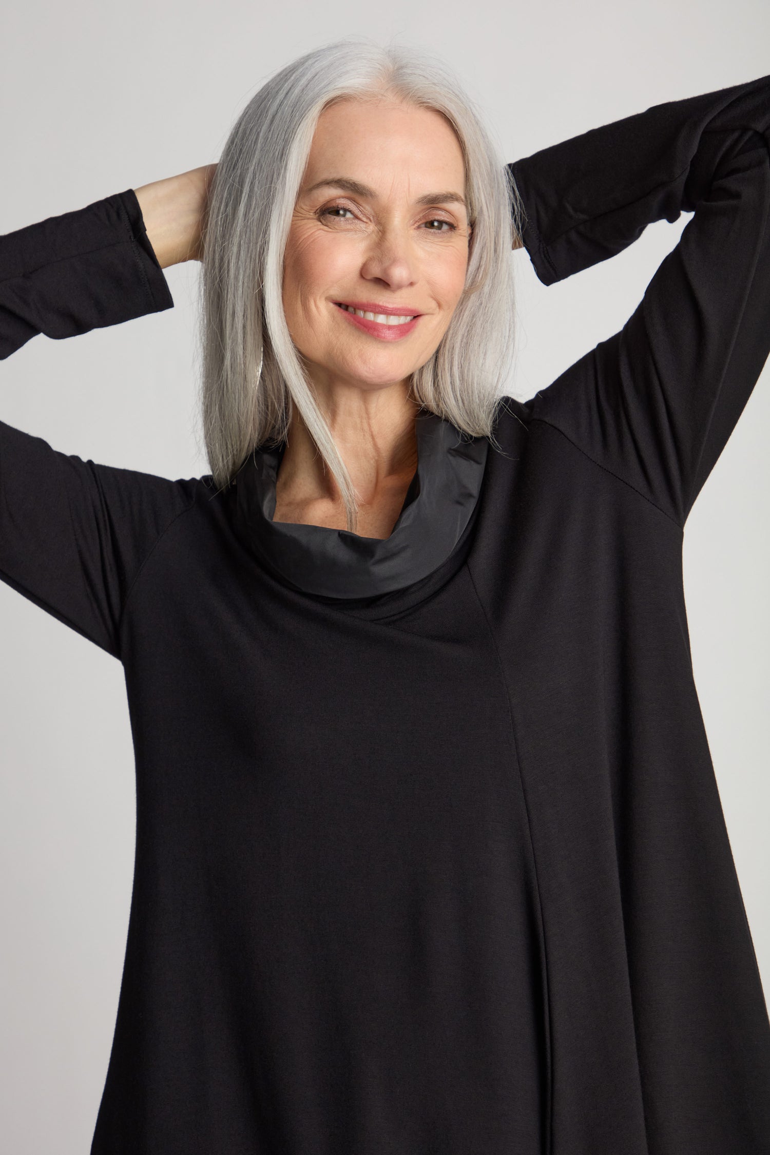 A woman with long white hair smiles while posing with her hands behind her head. She is wearing a black Cut Out Cowl Neck Dress, making for a chic statement piece.