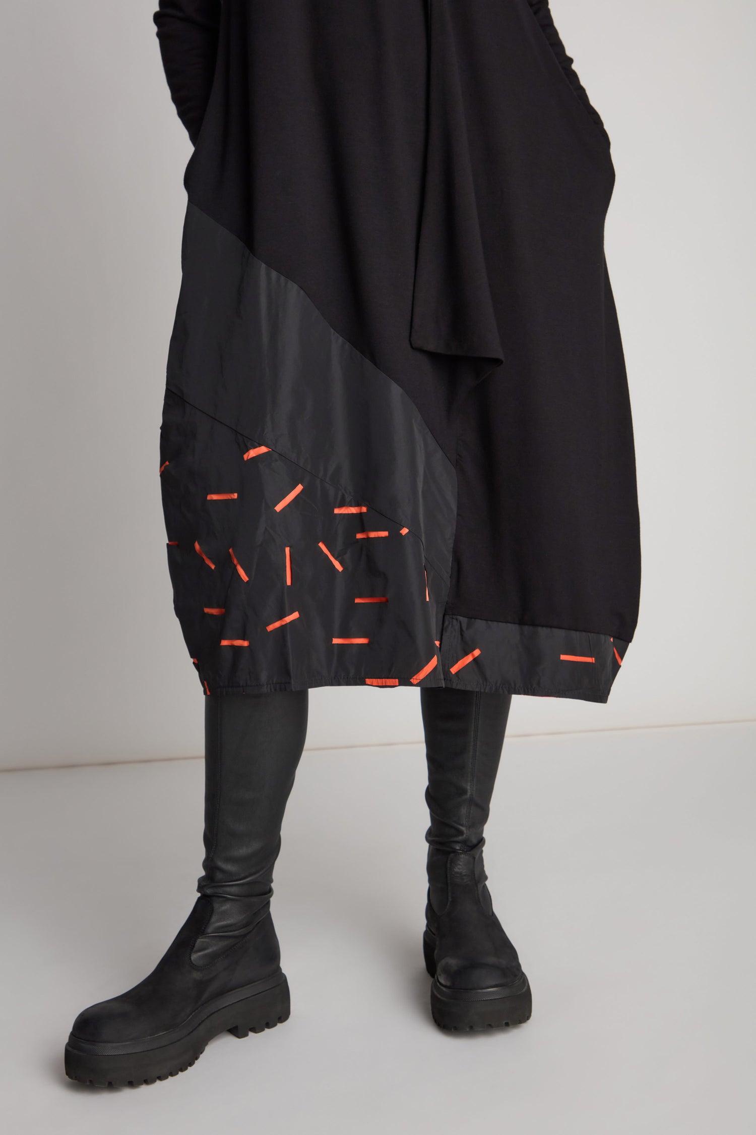 A person is wearing black clothing and chunky black boots. The Cut Out Cowl Neck Dress features a dark, patch pattern with orange accents near the hem, making it a true statement piece. The background is plain and light-colored.