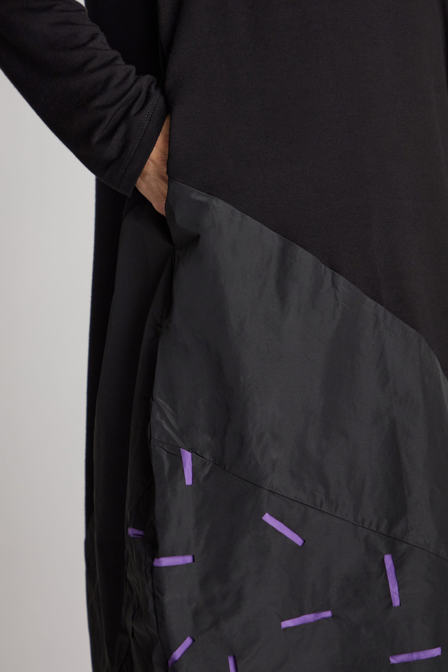 Close-up of a person wearing Luukaa's Cut Out Cowl Neck Dress in black, with their hand elegantly placed in a pocket. The smooth fabric features small purple rectangular accents that add a distinctive flair, making this dress a true statement piece.