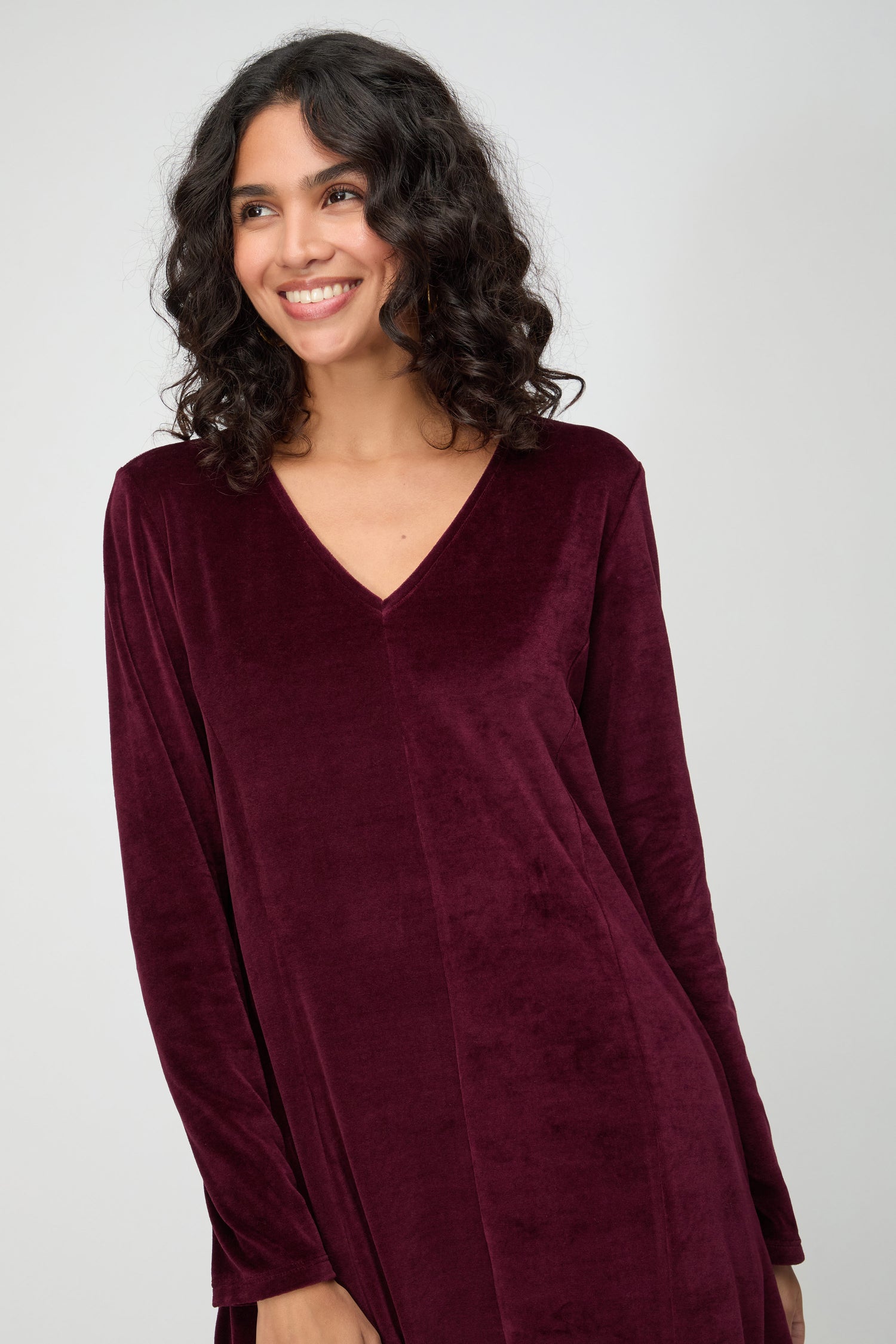 A woman with curly hair smiles while wearing the V Neck Velvet Jersey Bubble Dress in burgundy, featuring long sleeves and a V-neck, standing against a plain light background.