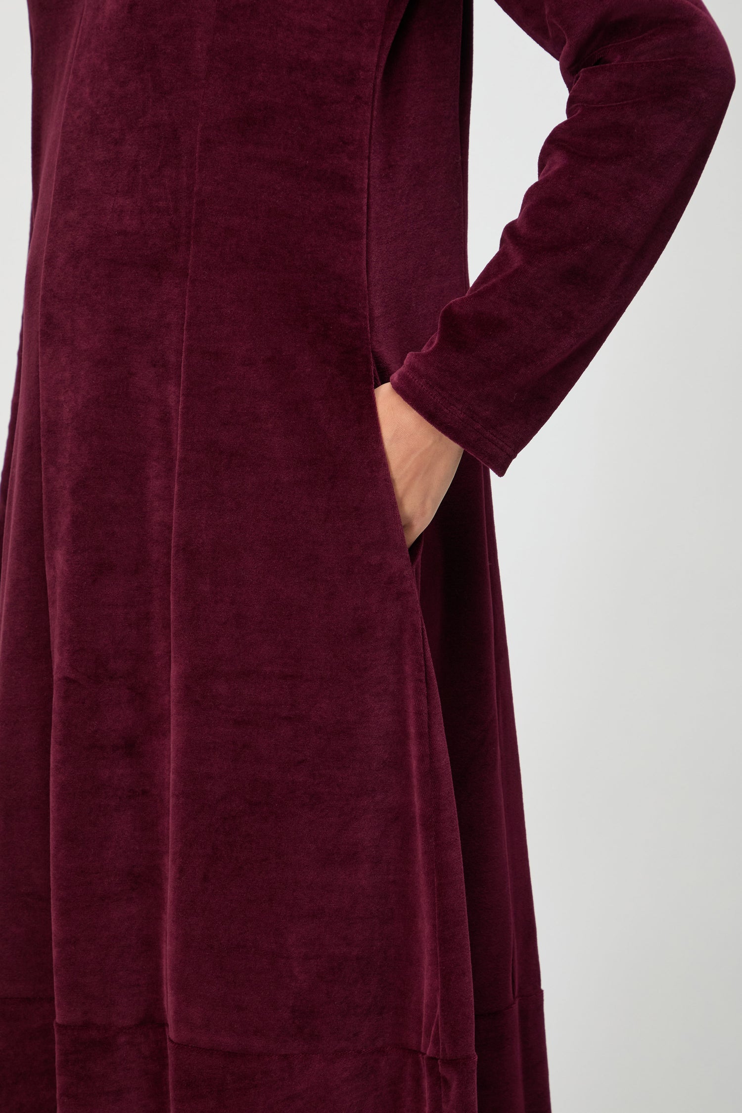 Person wearing the V Neck Velvet Jersey Bubble Dress in burgundy, featuring long sleeves and velvet jersey fabric, with their right hand in the pocket. Shown from shoulder to thigh.