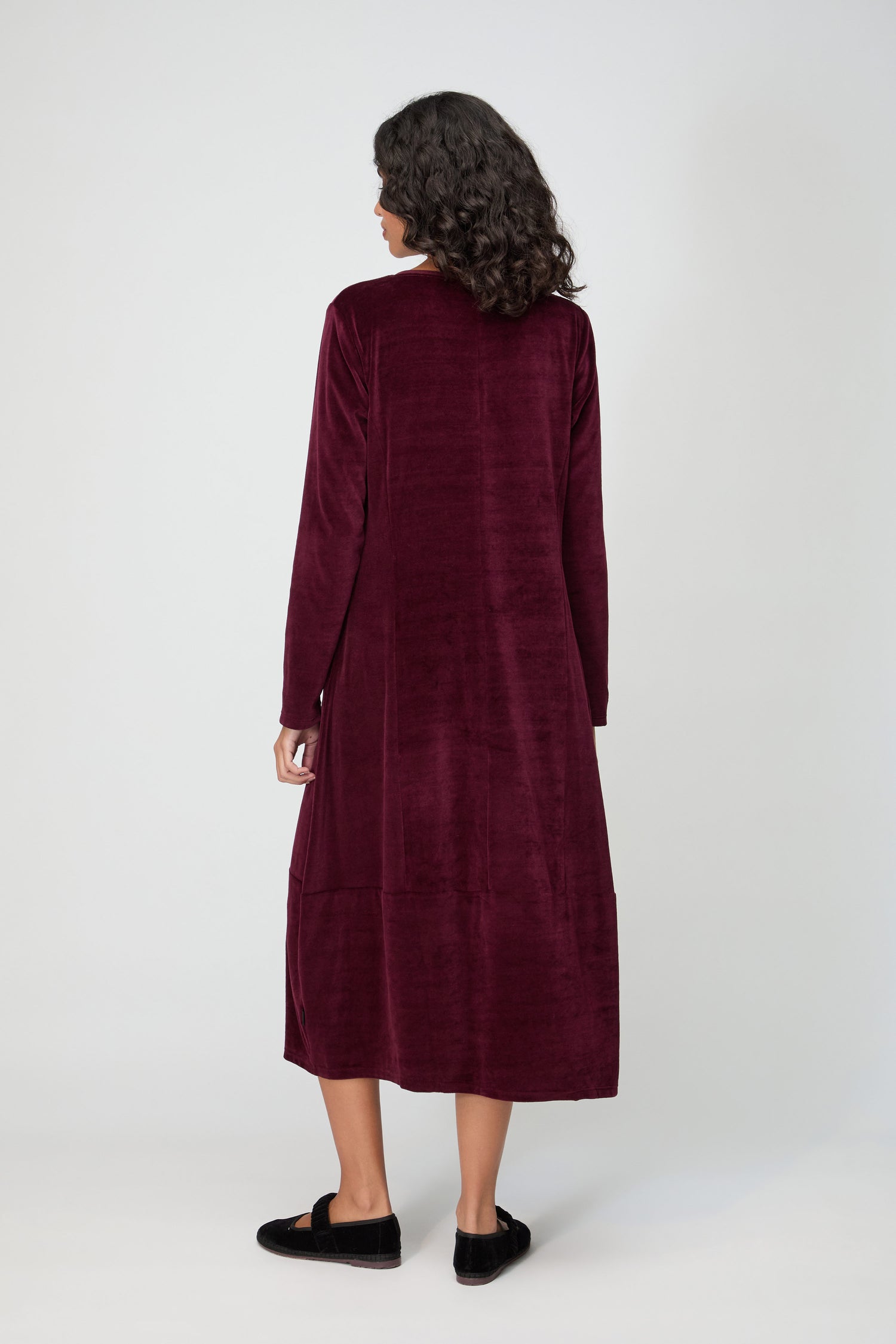 A person with medium-length curly hair is standing and facing away, wearing a long-sleeved, ankle-length V Neck Velvet Jersey Bubble Dress and black shoes against a plain background.