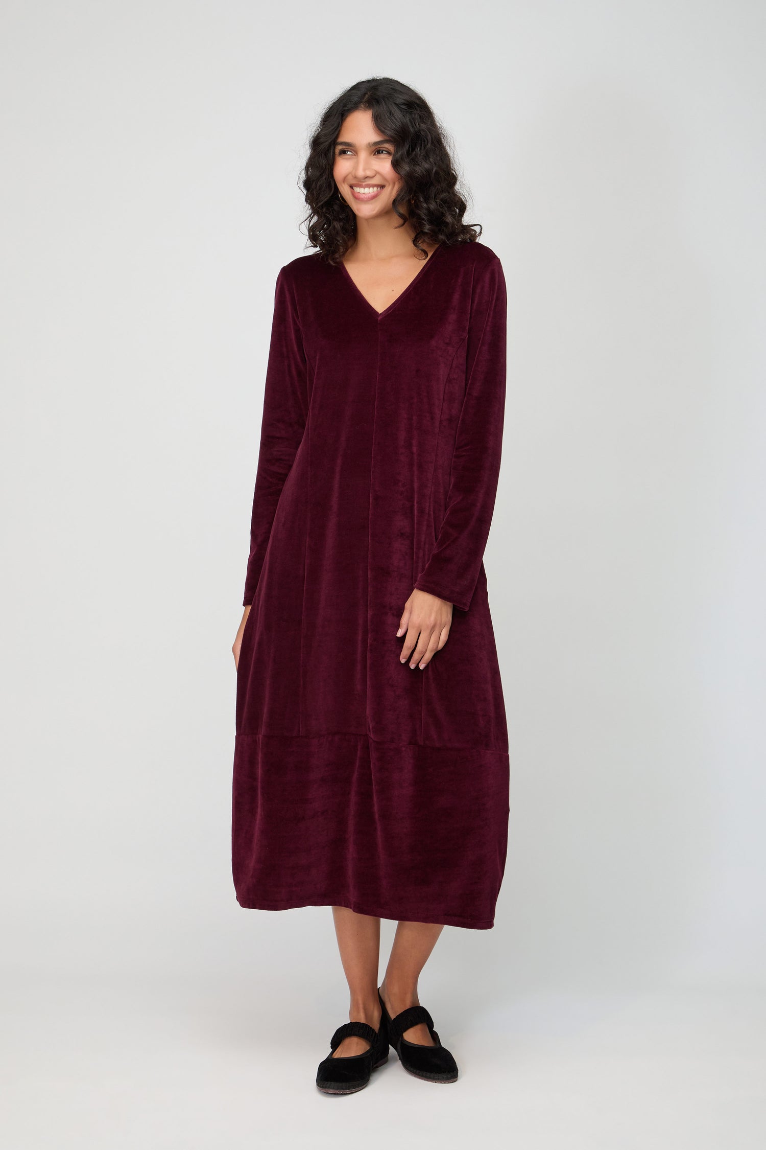 A person stands against a plain background, smiling with hands by their sides, wearing a long-sleeved burgundy V Neck Velvet Jersey Bubble Dress and black flat shoes.