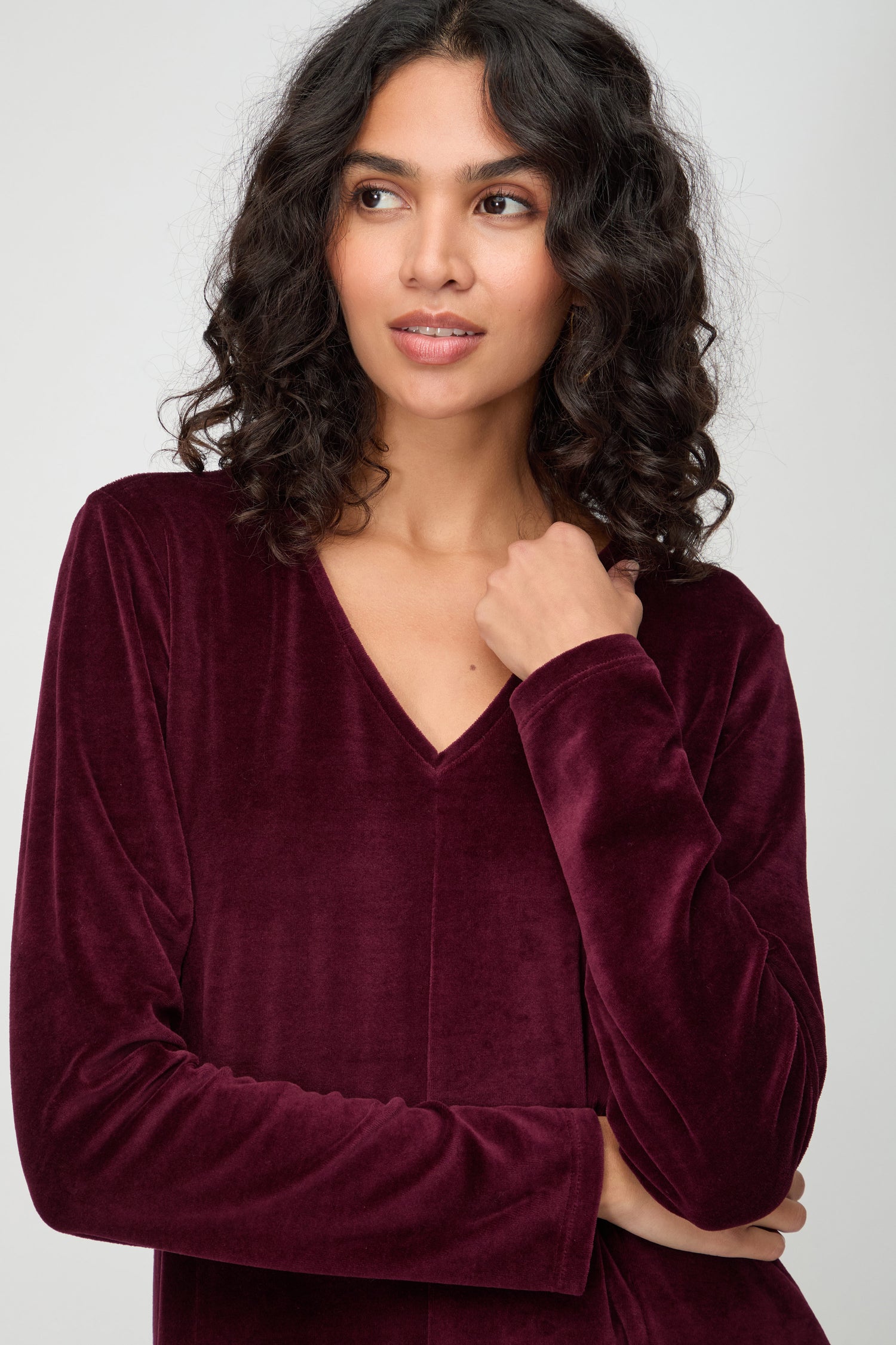 A woman with curly dark hair wears a V Neck Velvet Jersey Bubble Dress in burgundy and gazes off to the side against a plain background.
