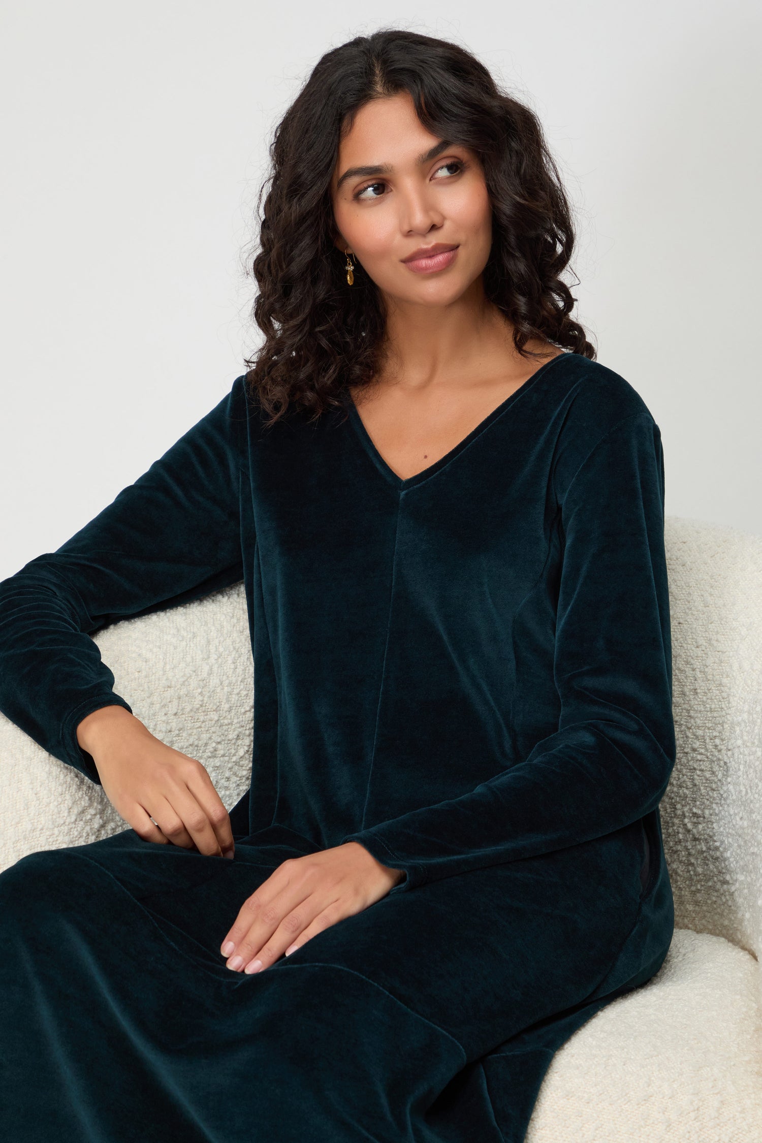 Woman with dark curly hair sits on a light textured chair, wearing a long-sleeve V Neck Velvet Jersey Bubble Dress and looking slightly to the side.