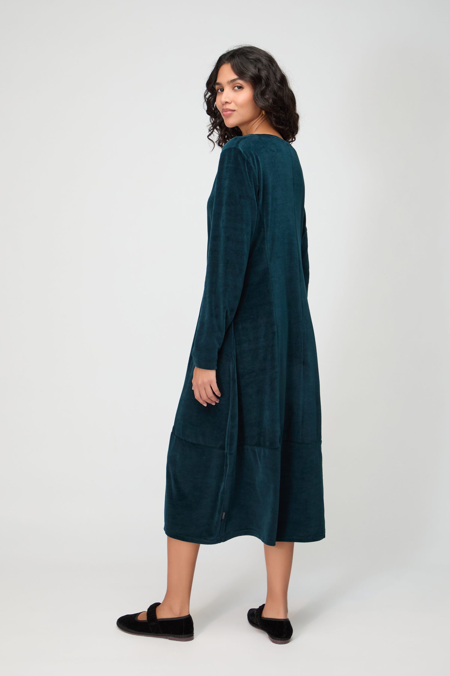 A person in a long-sleeved teal V Neck Velvet Jersey Bubble Dress and black slippers stands facing away, looking over their shoulder against a plain light background.