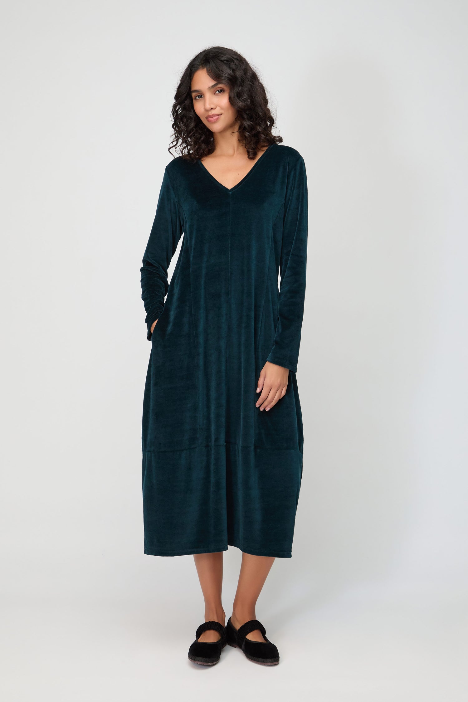 A woman with curly hair wears the V Neck Velvet Jersey Bubble Dress in dark teal with long sleeves and black shoes, standing against a plain light background.