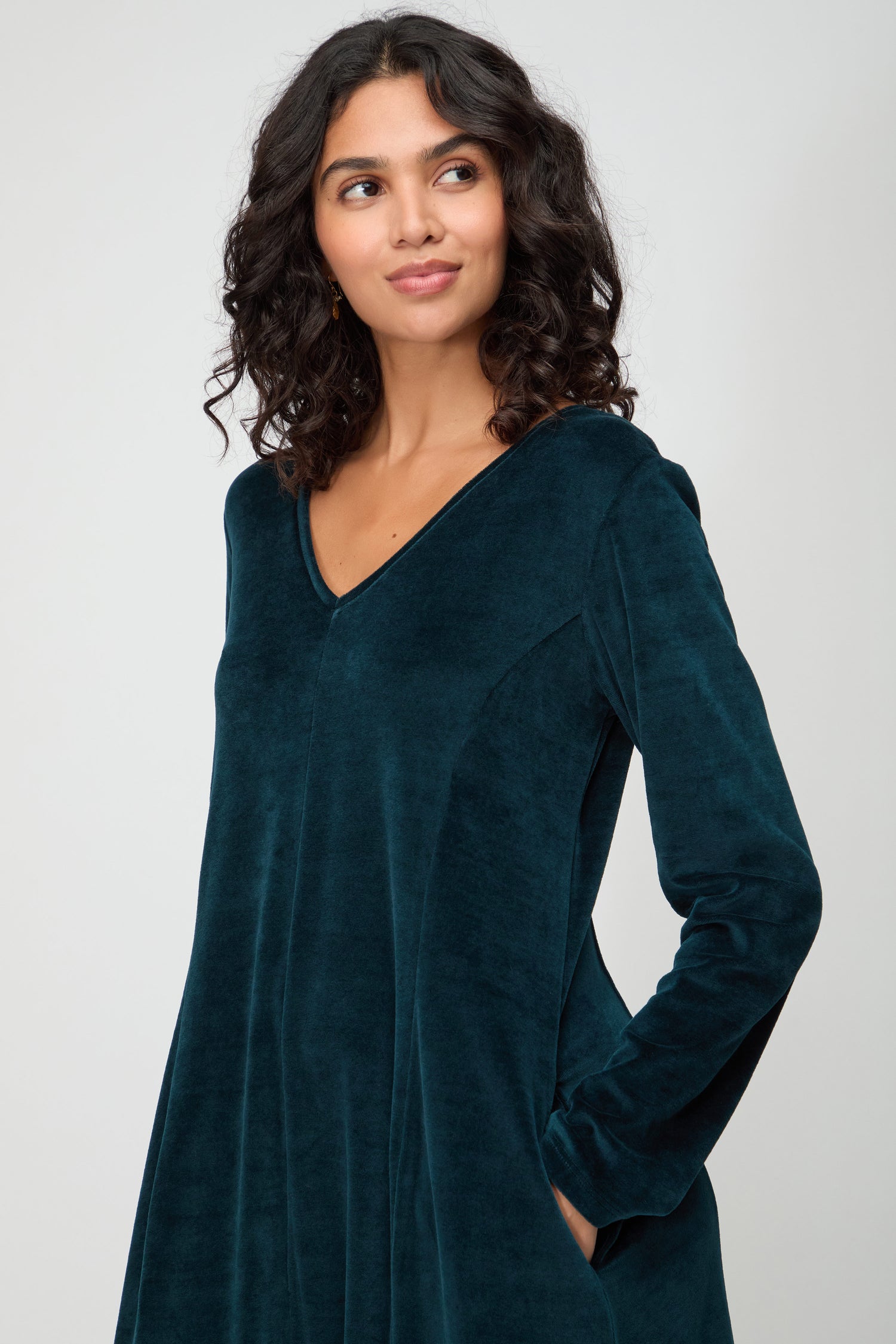 Woman with curly dark hair wears the V Neck Velvet Jersey Bubble Dress with long sleeves, standing against a plain light background and looking slightly to the side.