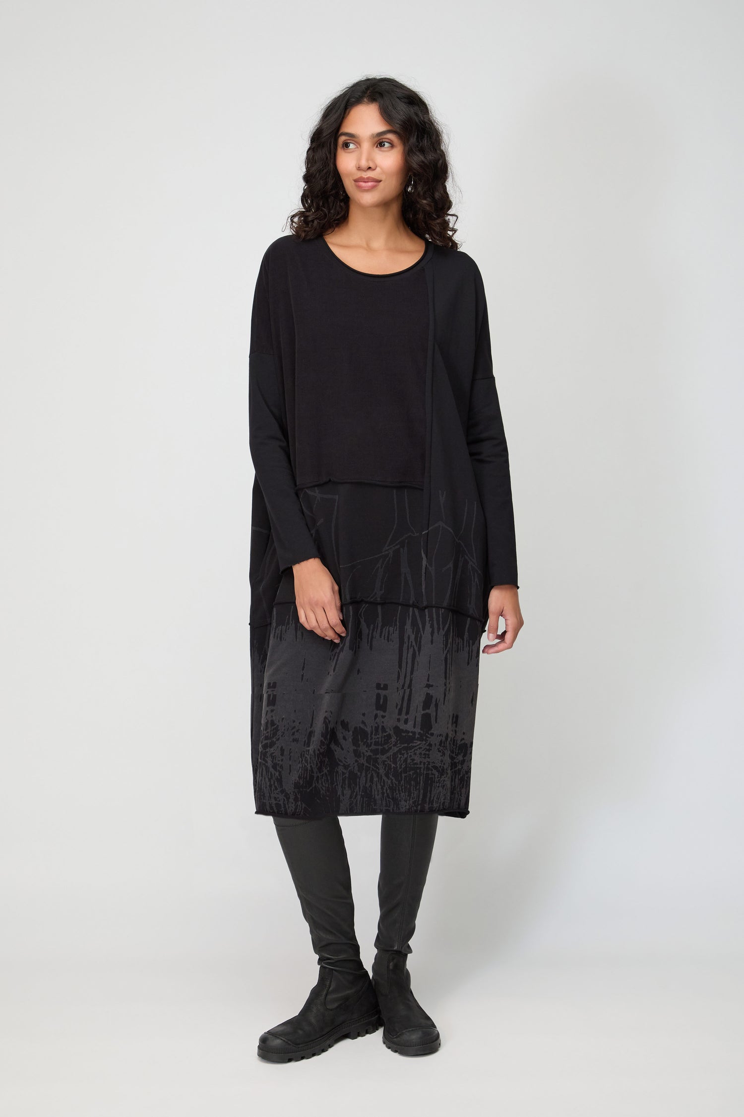 A woman stands against a plain background wearing the Burn Out Hem Detail Jersey Dress in black, featuring a layered effect and abstract patterns, paired with black leggings and black shoes.