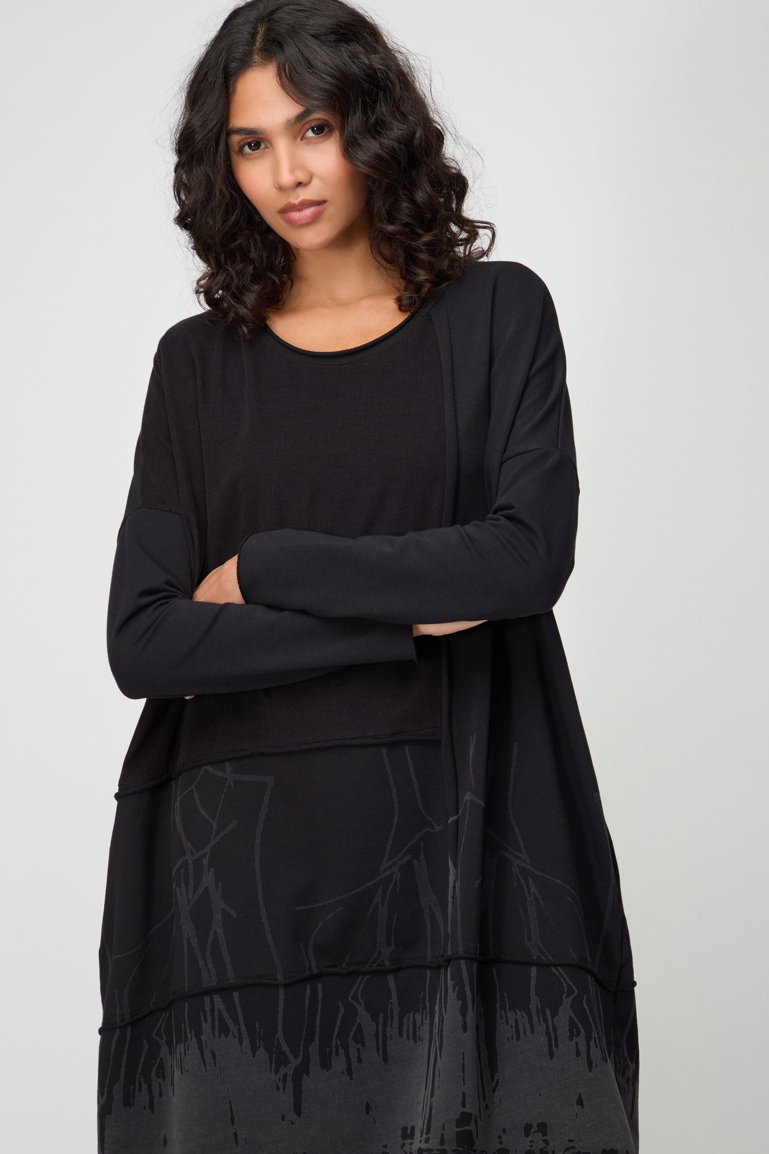 A woman with medium curly hair stands with arms crossed, wearing the Burn Out Hem Detail Jersey Dress with long sleeves and an abstract gray burn-out hem, set against a plain background.
