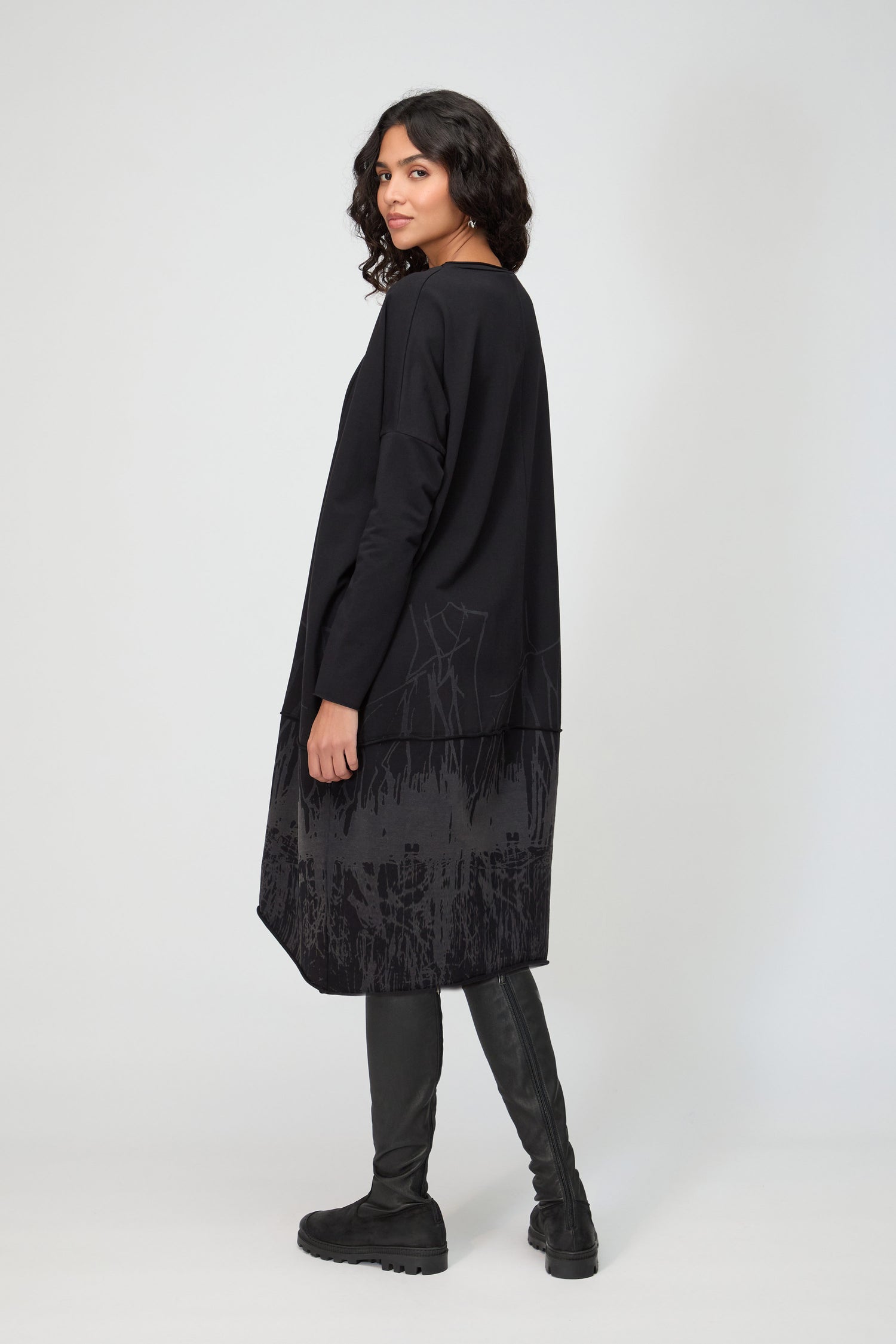 A woman stands sideways in the Burn Out Hem Detail Jersey Dress, featuring a burn-out hem and dark patterns, styled with black knee-high boots against a light background.