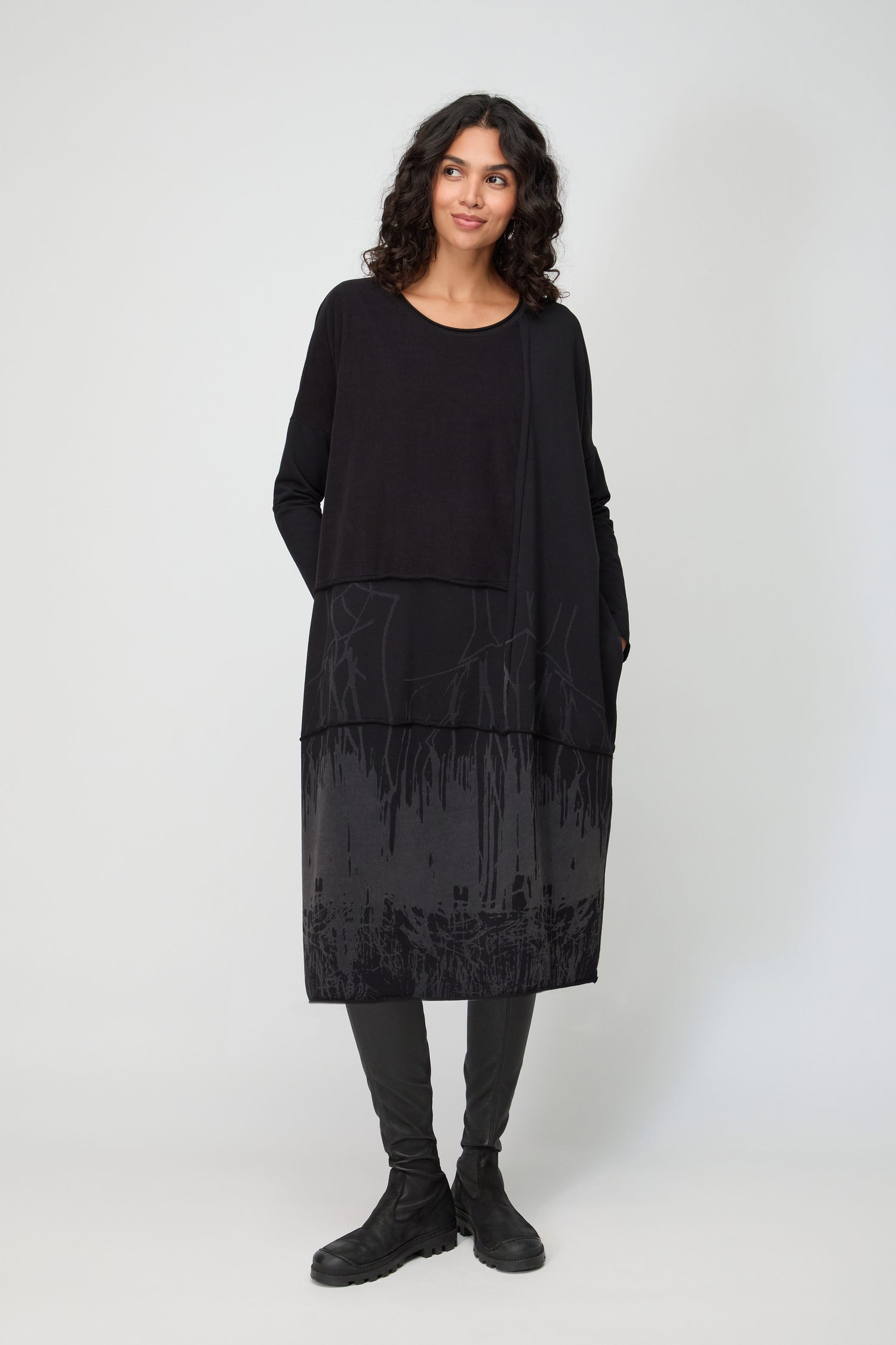 A person with curly hair stands against a plain background, wearing the Burn Out Hem Detail Jersey Dress featuring a layered effect and abstract patterns, paired with black leggings and boots.