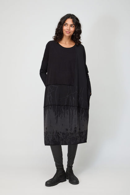 A person with curly hair stands against a plain background, wearing the Burn Out Hem Detail Jersey Dress featuring a layered effect and abstract patterns, paired with black leggings and boots.