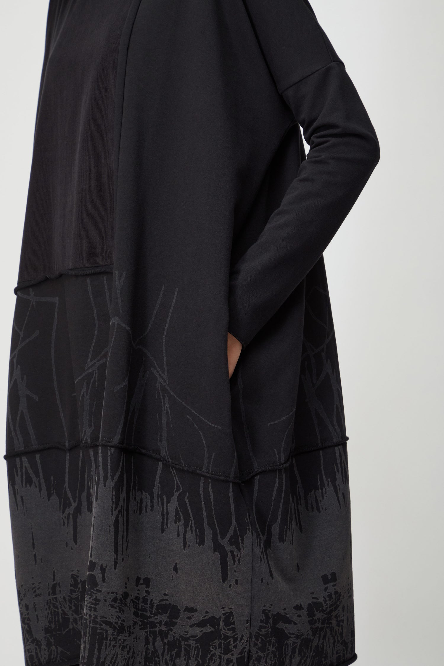 Model wearing the Burn Out Hem Detail Jersey Dress with long sleeves, gray abstract patterns, and a burn-out hem, shown from shoulders down with one hand in a pocket.