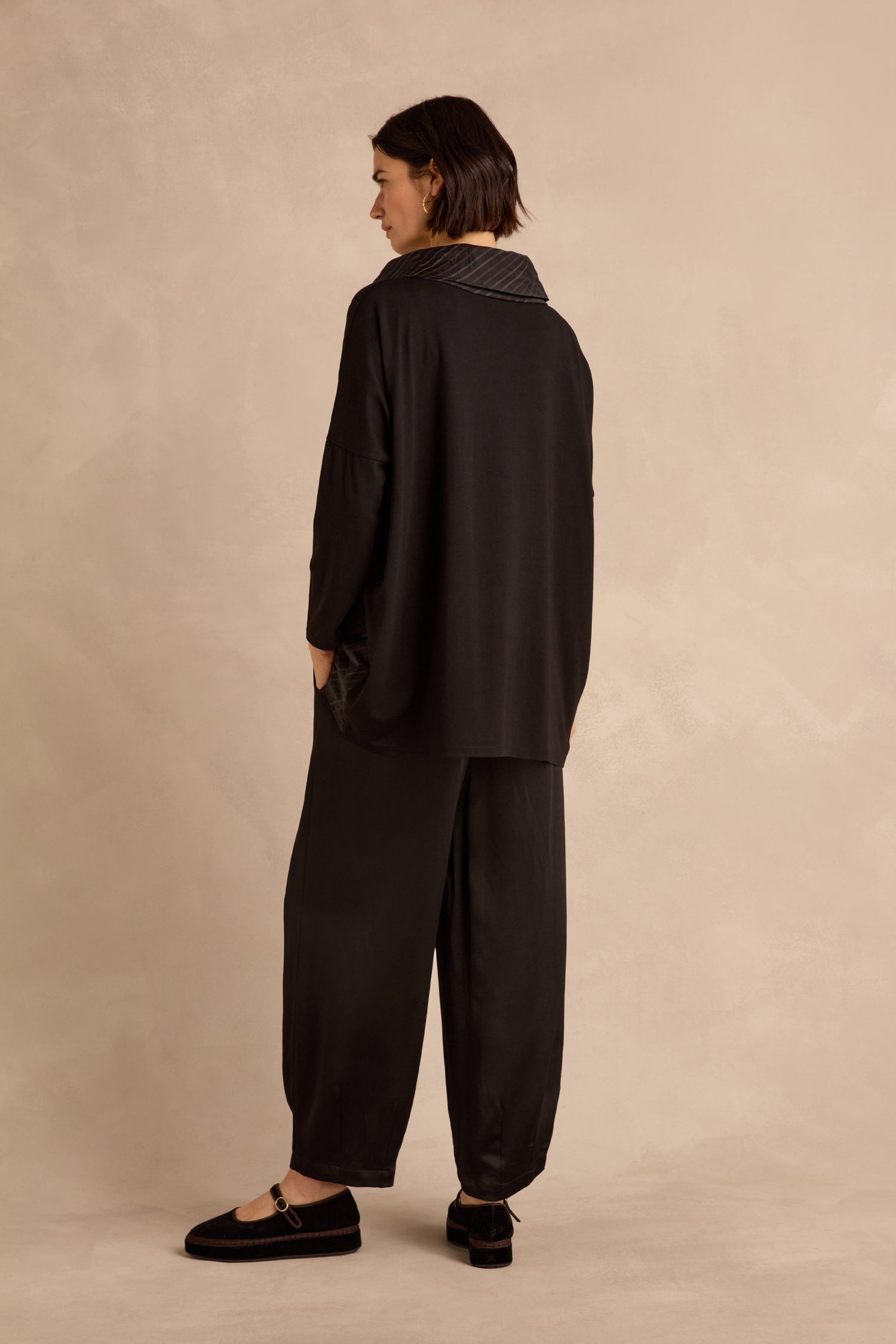 Person standing with back turned, wearing a loose black Stripe Jersey Taffeta Tunic and black shoes, against a plain beige background.
