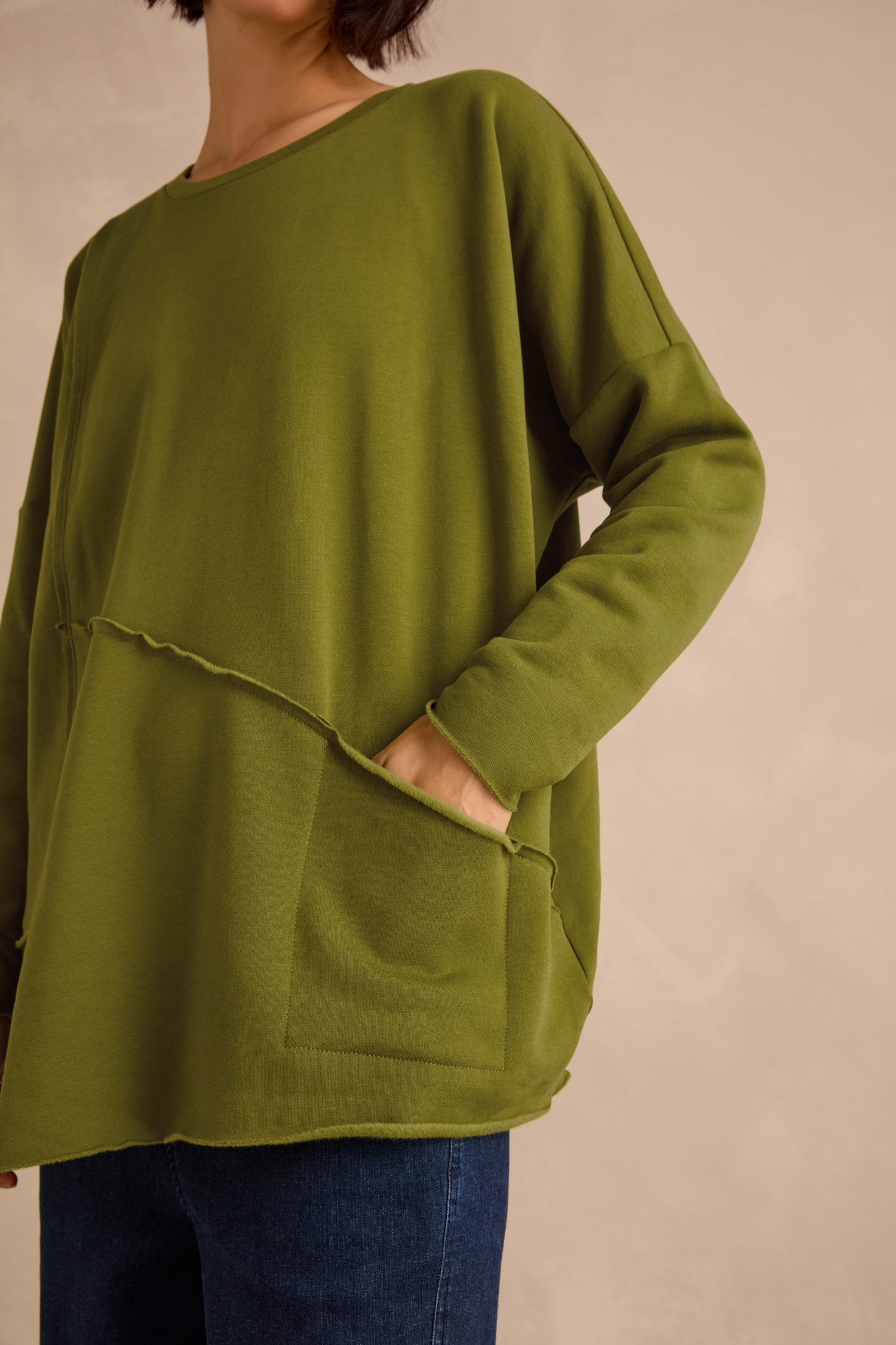 A person wearing the Seam Detail Pocket Top—a relaxed, oversized long-sleeve top in green with seam details and a front pocket, made from soft cotton lyocell stretch jersey—stands against a neutral background with one hand in the pocket, paired with blue jeans.