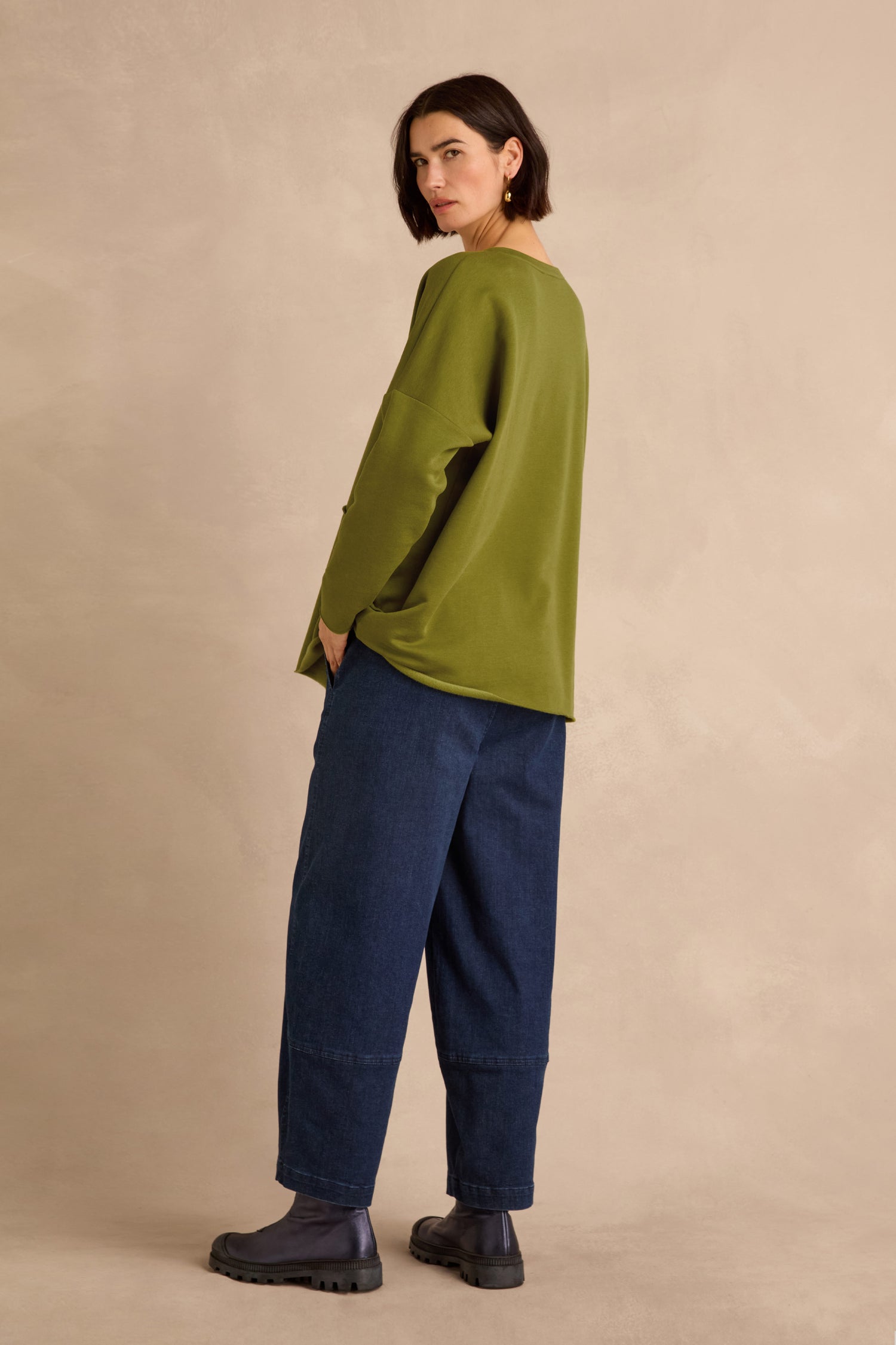A person stands sideways wearing the Seam Detail Pocket Top in olive green, crafted from cotton lyocell stretch jersey, paired with wide-leg dark blue jeans and black boots against a beige background.