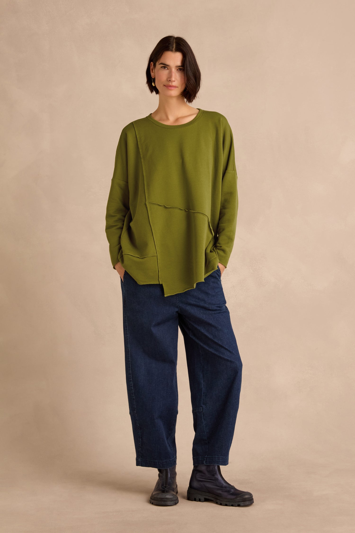 A person stands against a beige background wearing a loose green Seam Detail Pocket Top, dark blue wide-leg jeans, and black boots, with hands in pockets and short dark hair.