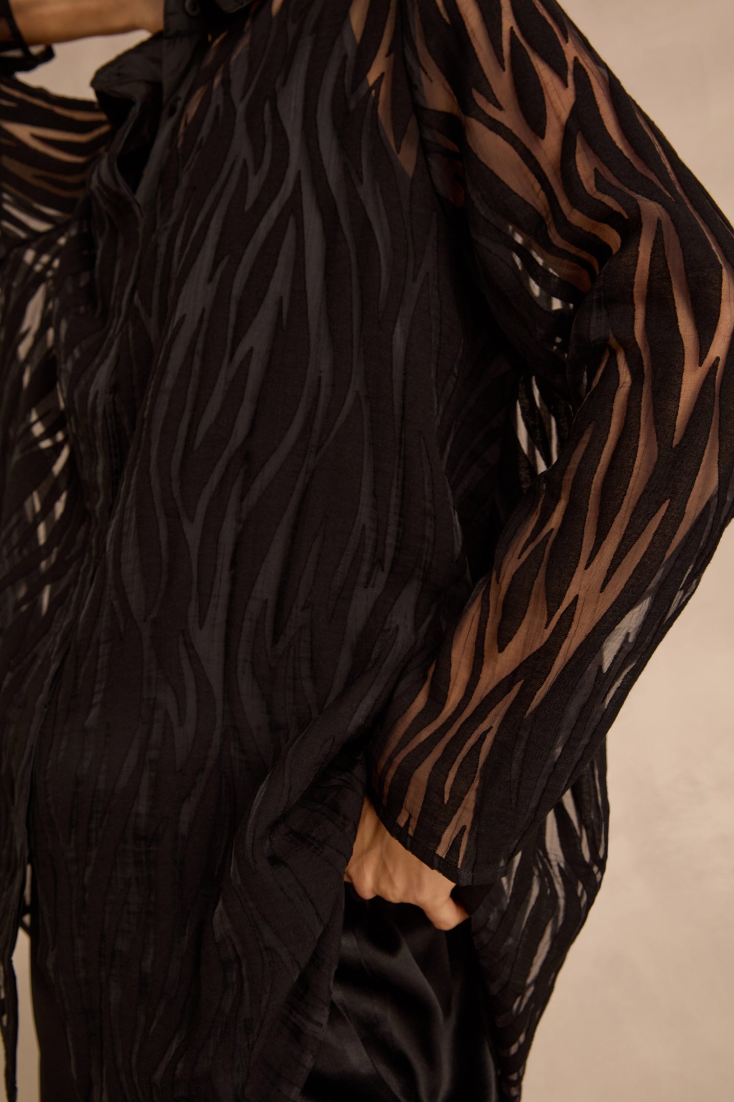A person wearing the Wave Cut Out Shirt, a sheer black top featuring a wavy, zebra-like pattern, with their hand in a pocket.