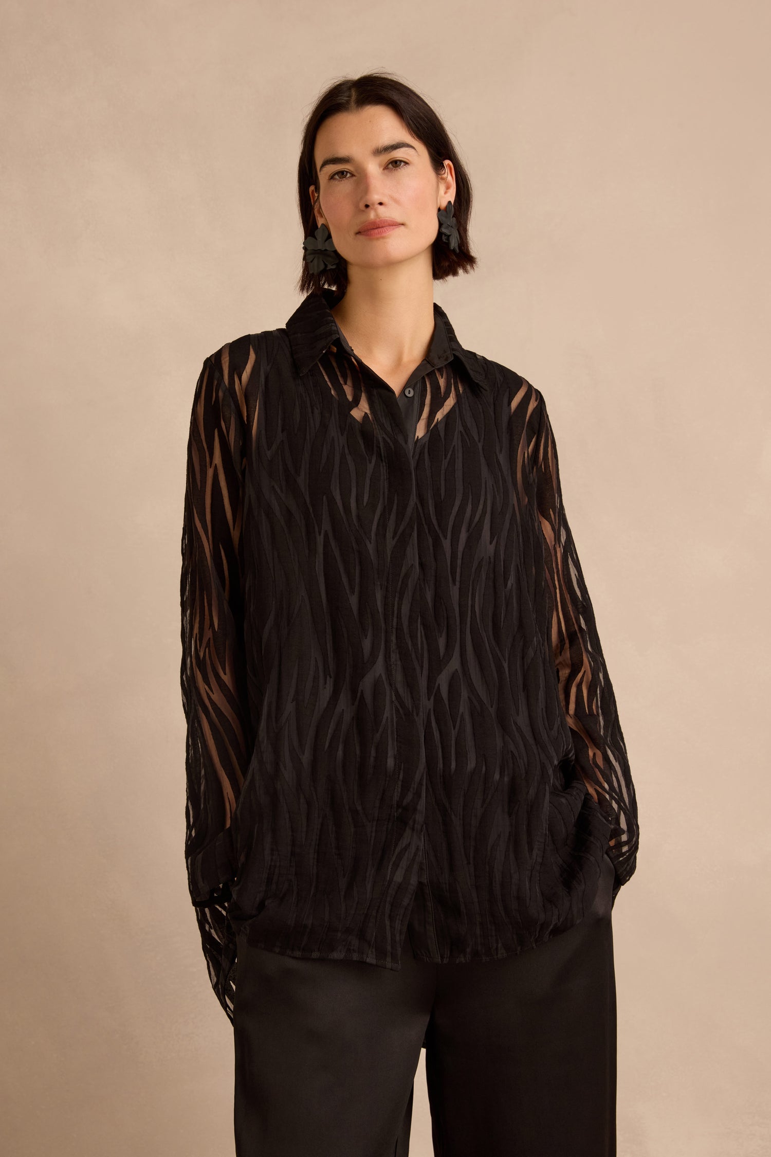 A woman wears the Wave Cut Out Shirt, featuring sheer black viscose-blend fabric with patterned wave cut outs, paired with black pants. The long sleeves and tunic length of the shirt add an elegant, modern touch as she stands against a plain beige background.