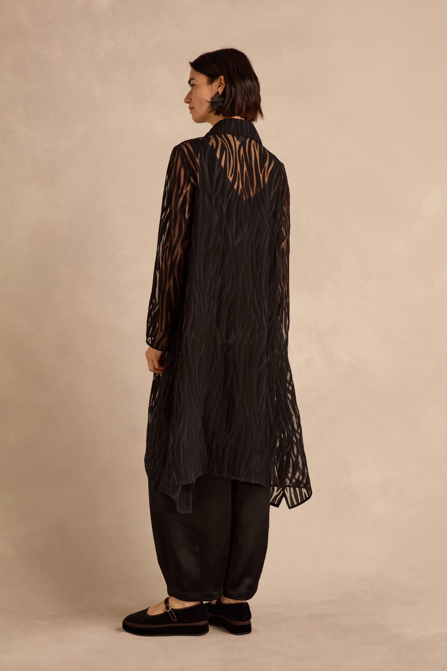 A person stands facing away against a neutral background, wearing the Wave Cut Out Long Shirt—sheer with a wave cutout—paired with black pants and black shoes.
