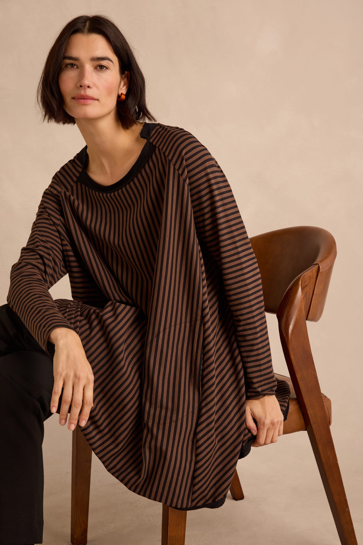 A woman with short dark hair sits on a wooden chair, wearing the Stripe Jersey Wide Neck Tunic and black pants, posing against a neutral background.