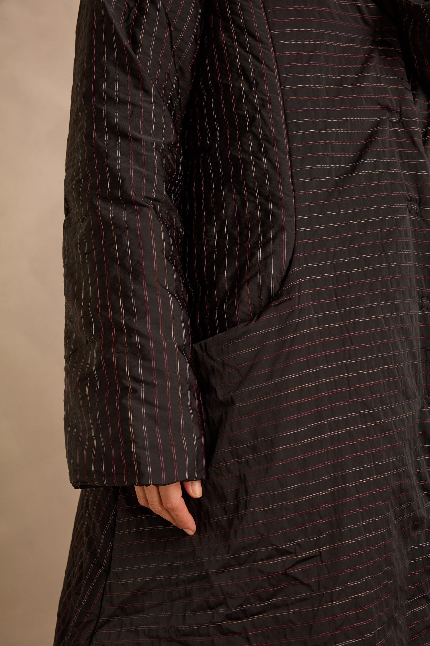 A person wearing a black Striped Puffer Coat by Luukaa with subtle red stripes, standing with one hand partially visible near a large pocket.