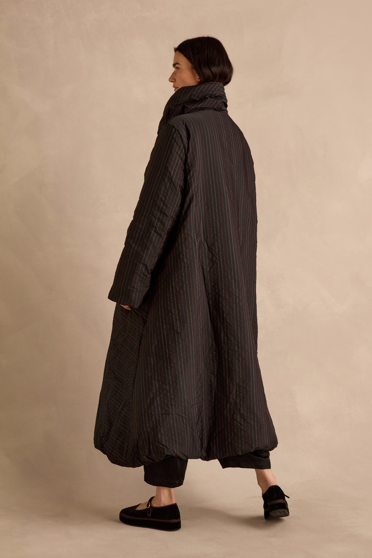 A person stands sideways against a neutral backdrop, wearing the Striped Puffer Coat by Luukaa—a long, dark quilted outerwear piece with vertical stripes—paired with cropped black pants and black loafers.