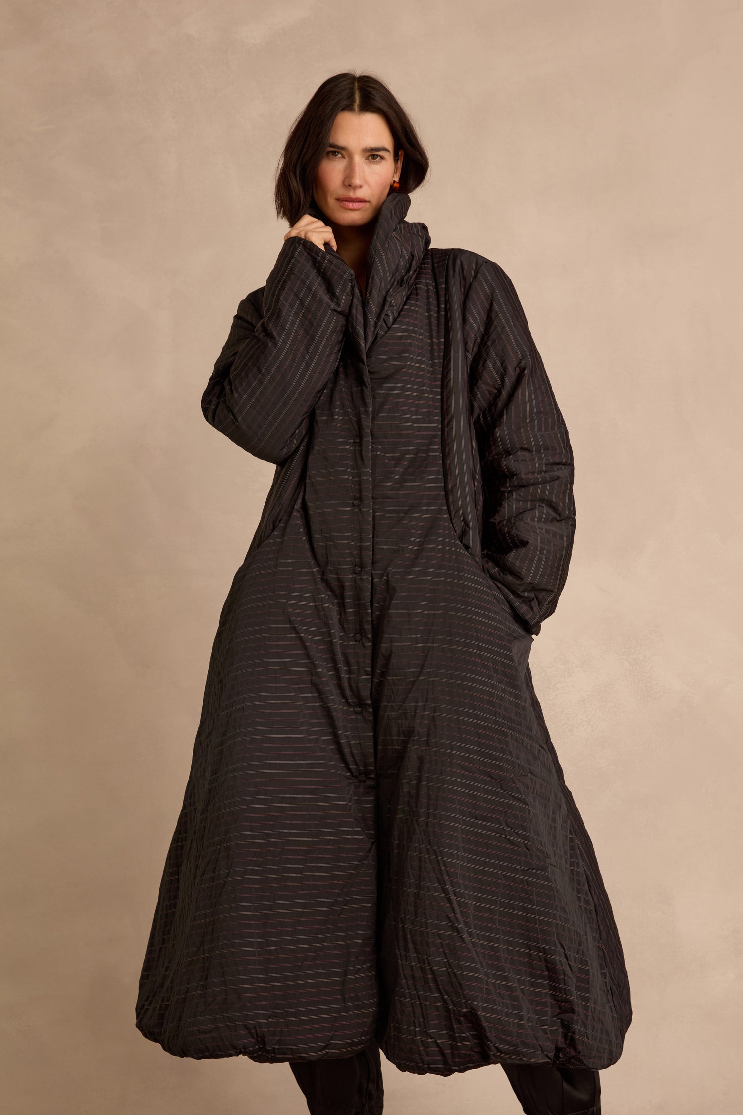 A person stands against a beige background wearing a long, black, oversized Striped Puffer Coat with their hands in the pockets.