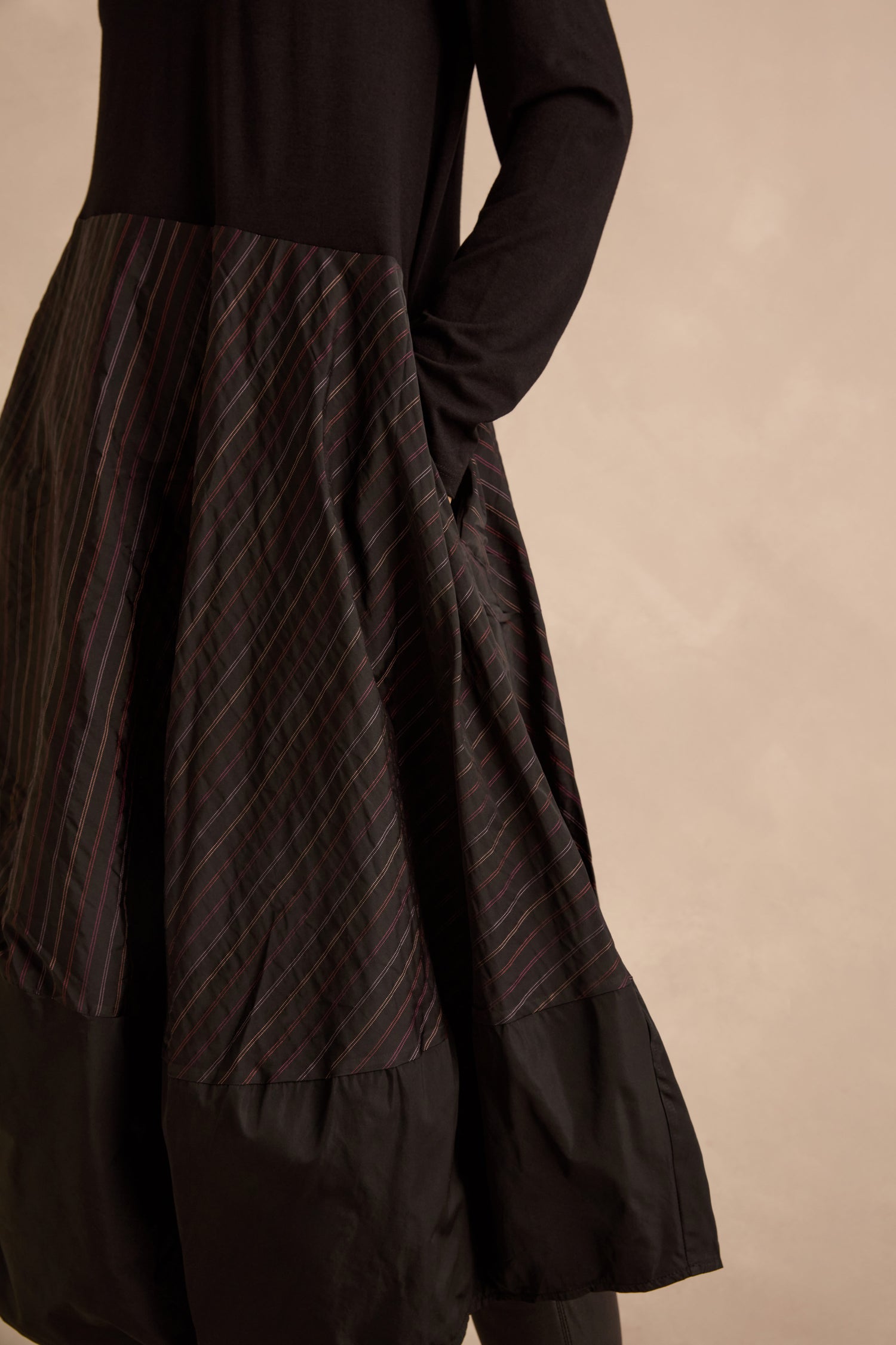 Person in a Stripe Jersey Taffeta Bubble Dress featuring black long sleeves, a flared patterned skirt, and one hand in a pocket, standing against a beige background.