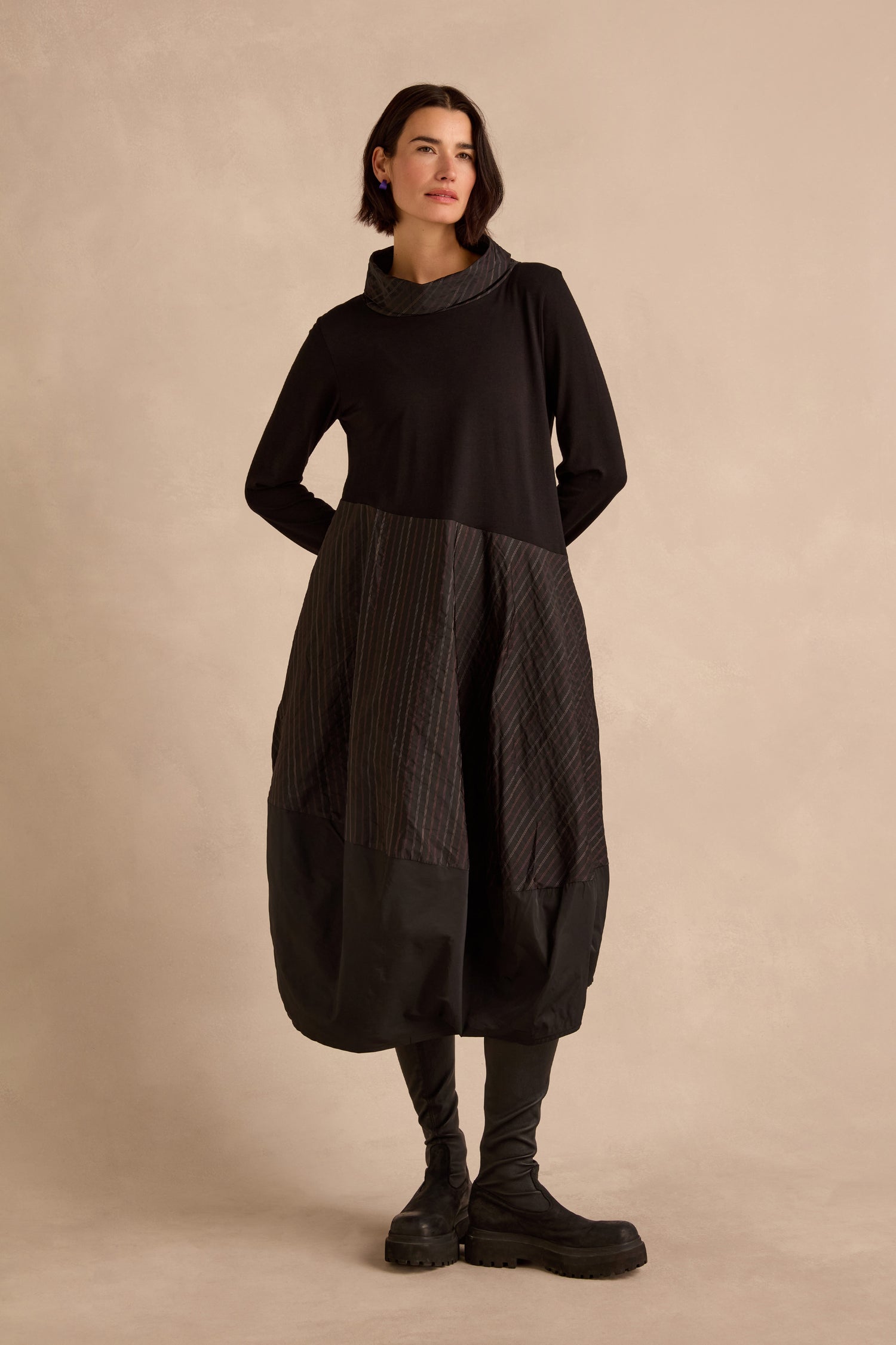 A woman stands against a beige background wearing the long-sleeved Stripe Jersey Taffeta Bubble Dress with a mixed fabric skirt and black boots.
