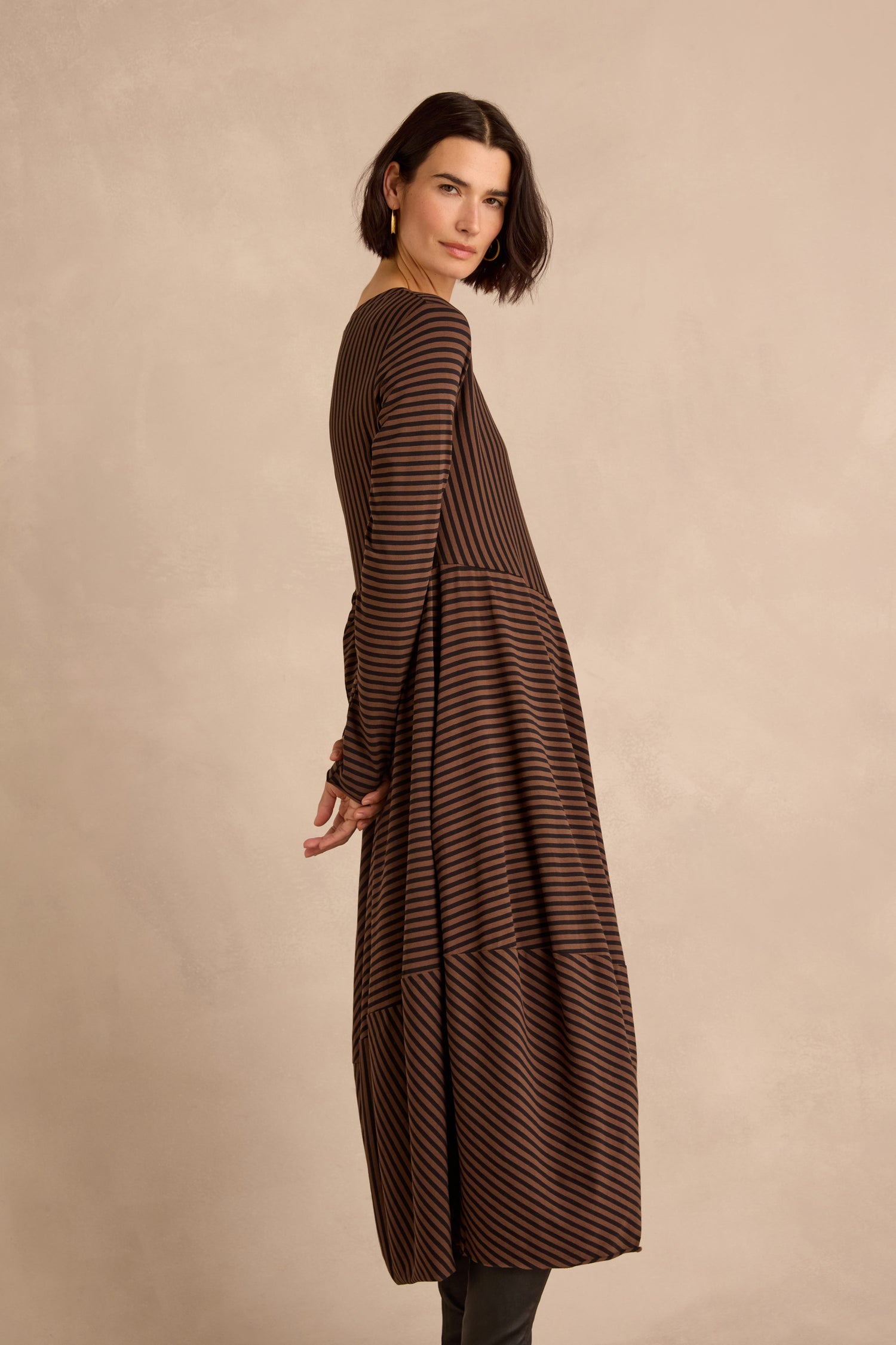 A woman with short dark hair stands sideways against a plain beige background, wearing the Draped Stripe Jersey Dress, a long dress featuring brown and black stripes.