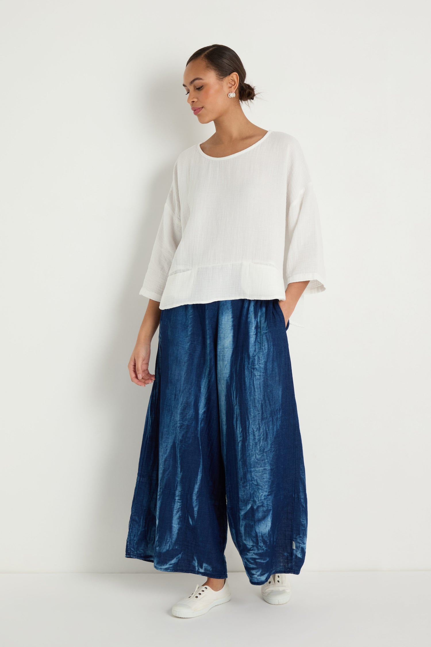 A woman stands against a white wall wearing the Crinkle Two-Pocket Top, wide-leg shiny blue pants, and white sneakers.